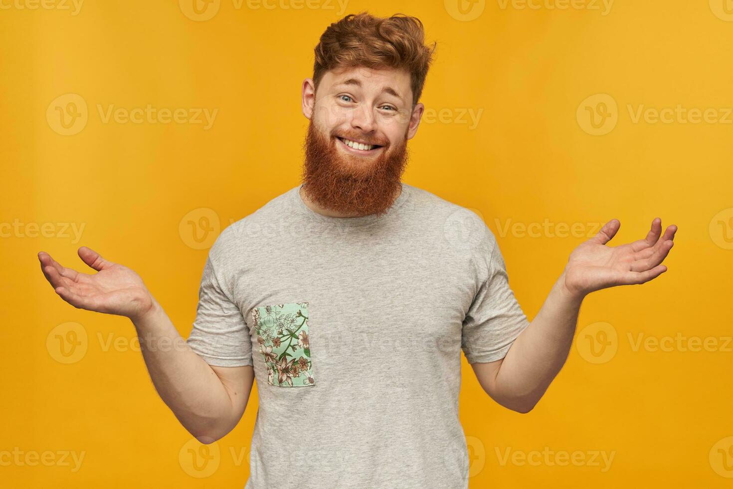 Indoor shot of young redhead male with big beard, holding copy space with both hands, smiling with confused, irritated facial expression. Isolated over yellow background photo