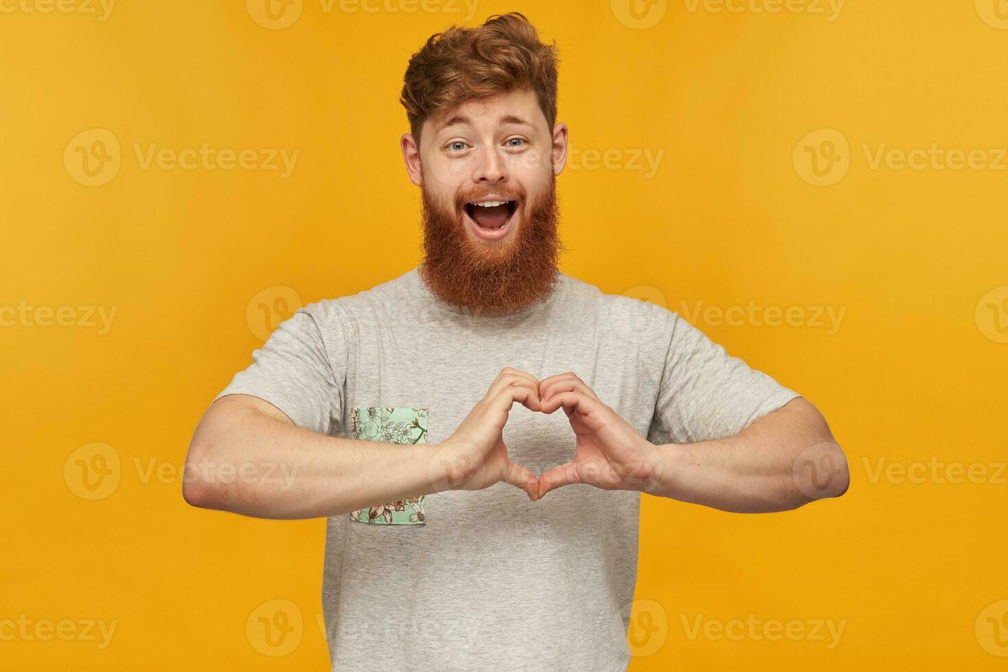 indoor shot of young bearded male, with red hair and big beard, shows heart sign with his hands, smiles broadly and feels happy, isolated over yellow background. photo