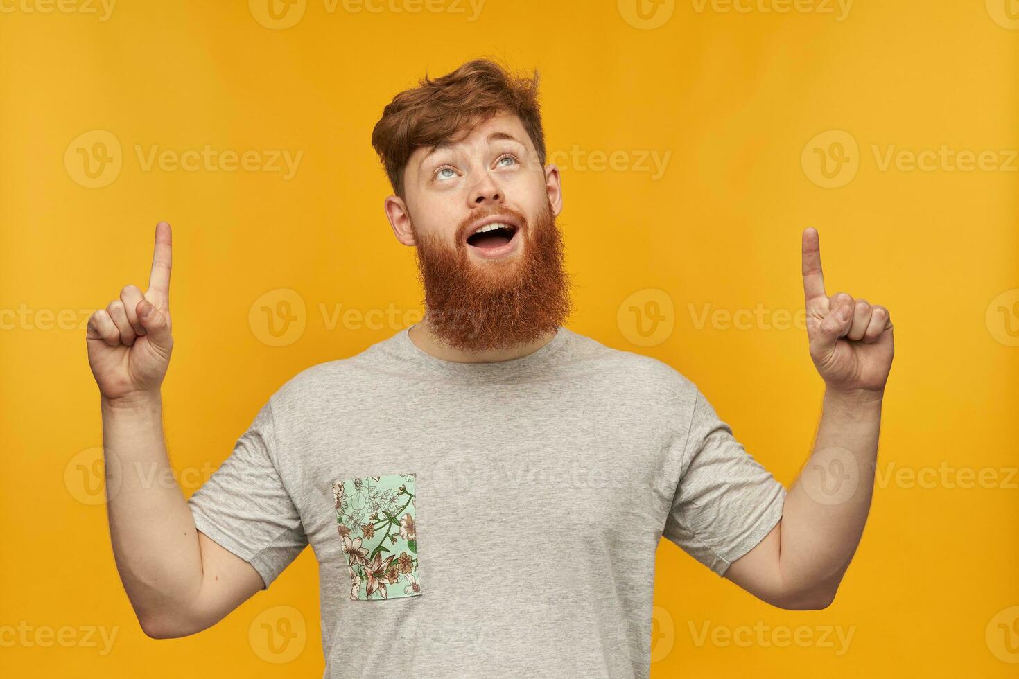 Indoor shot of young bearded male, wears grey t-shirt, pointing with both hands at copy space above him, smiling with amazed facial expression. Isolated over yellow background. photo