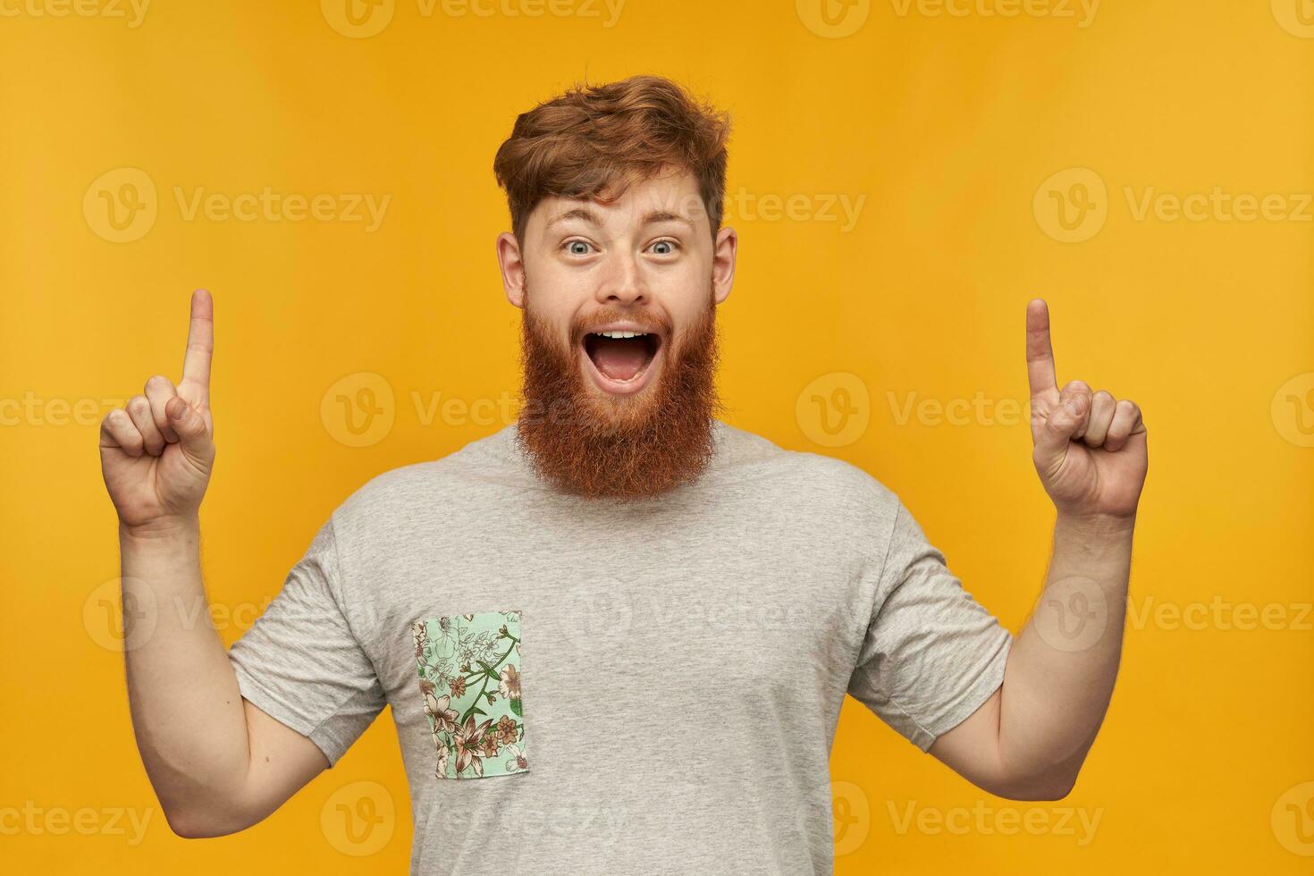 Indoor shot of young bearded male, wears grey t-shirt, pointing with both hands at copy space above him, smiling with amazed facial expression. Isolated over yellow background. photo