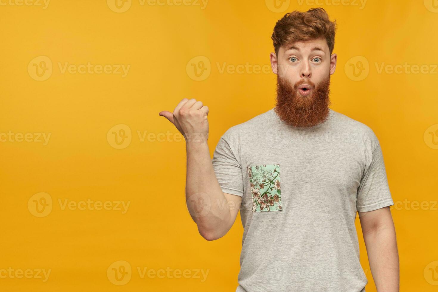 indoor shot of amazed, positive young bearded male with red hair, pointing with a finger left at copy space with amazed facial expression. Isolated over yellow background photo