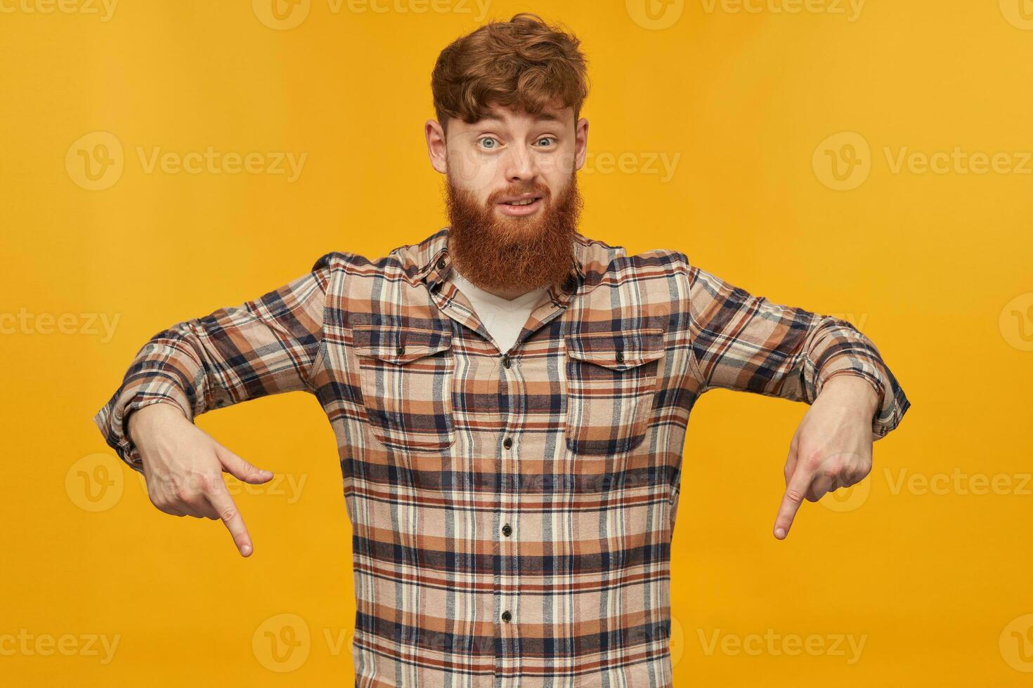 Young bearded male, wears casual shirt, with big beard and red hair, indicates with both hands down, isolated over yellow background photo