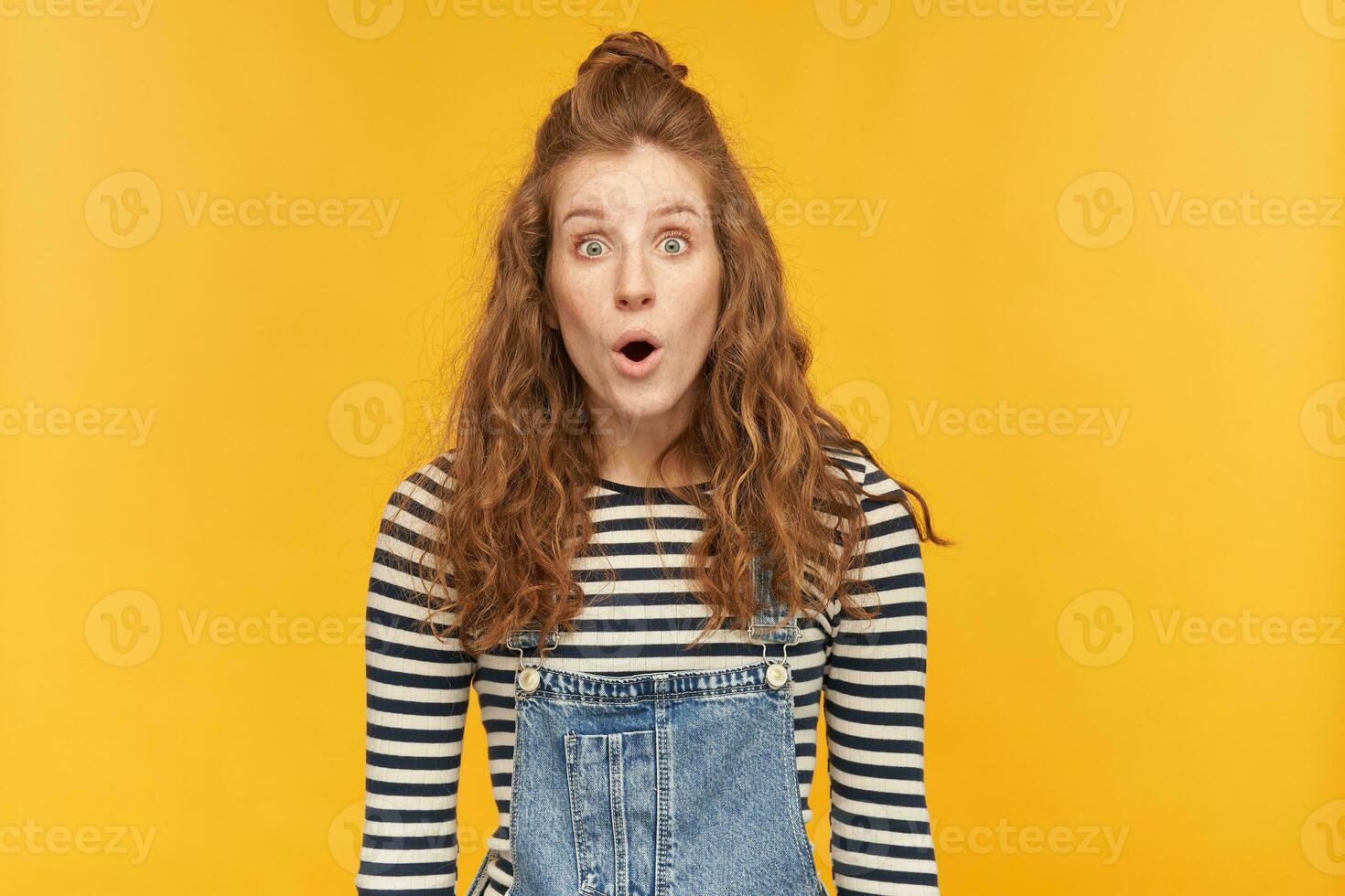 indoor portrait of young ginger curly female posing over yellow bright background looking into camera with shocked facial expression photo