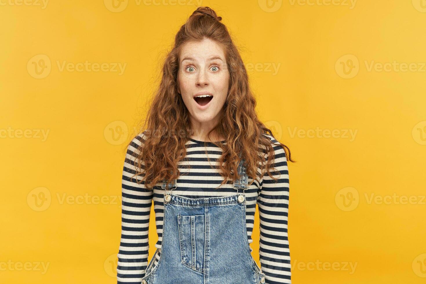 indoor portrait of amazed young ginger girl, wears denim overalls and stripped shirt, starring into camera with widely opened mouth and eyes. shocked, amazed facial expression. photo