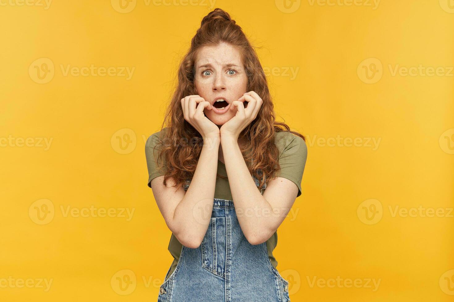 Indoor shot of young female with a long red hair, wears denim overalls and green t-shirt, starring into camera with scared facial expression while watching horror movie. photo