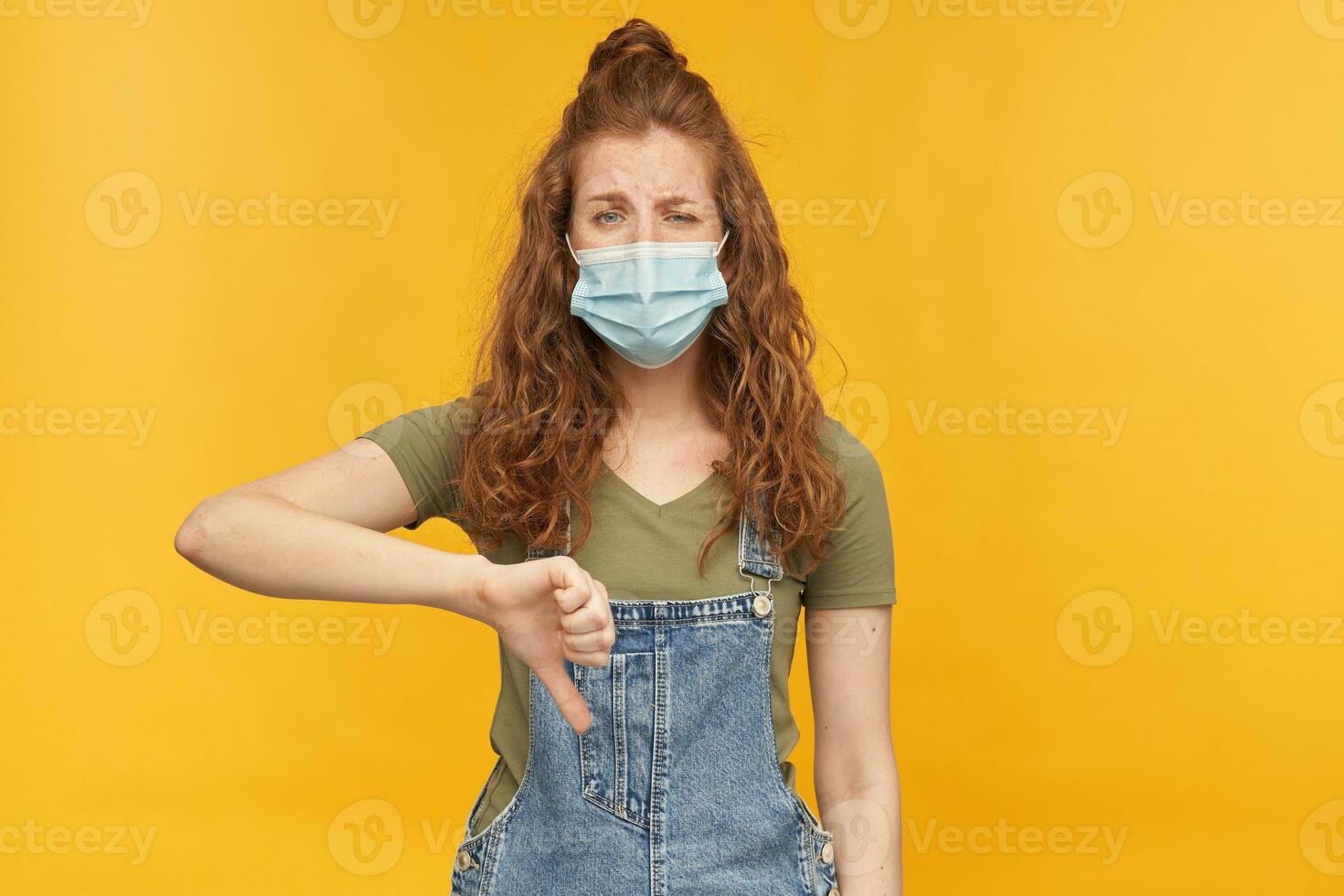 indoor studio shot of young ginger female with long red curly hair shows thumbs down gesture with sad facial expression, wears face mask. isolated over yellow background photo