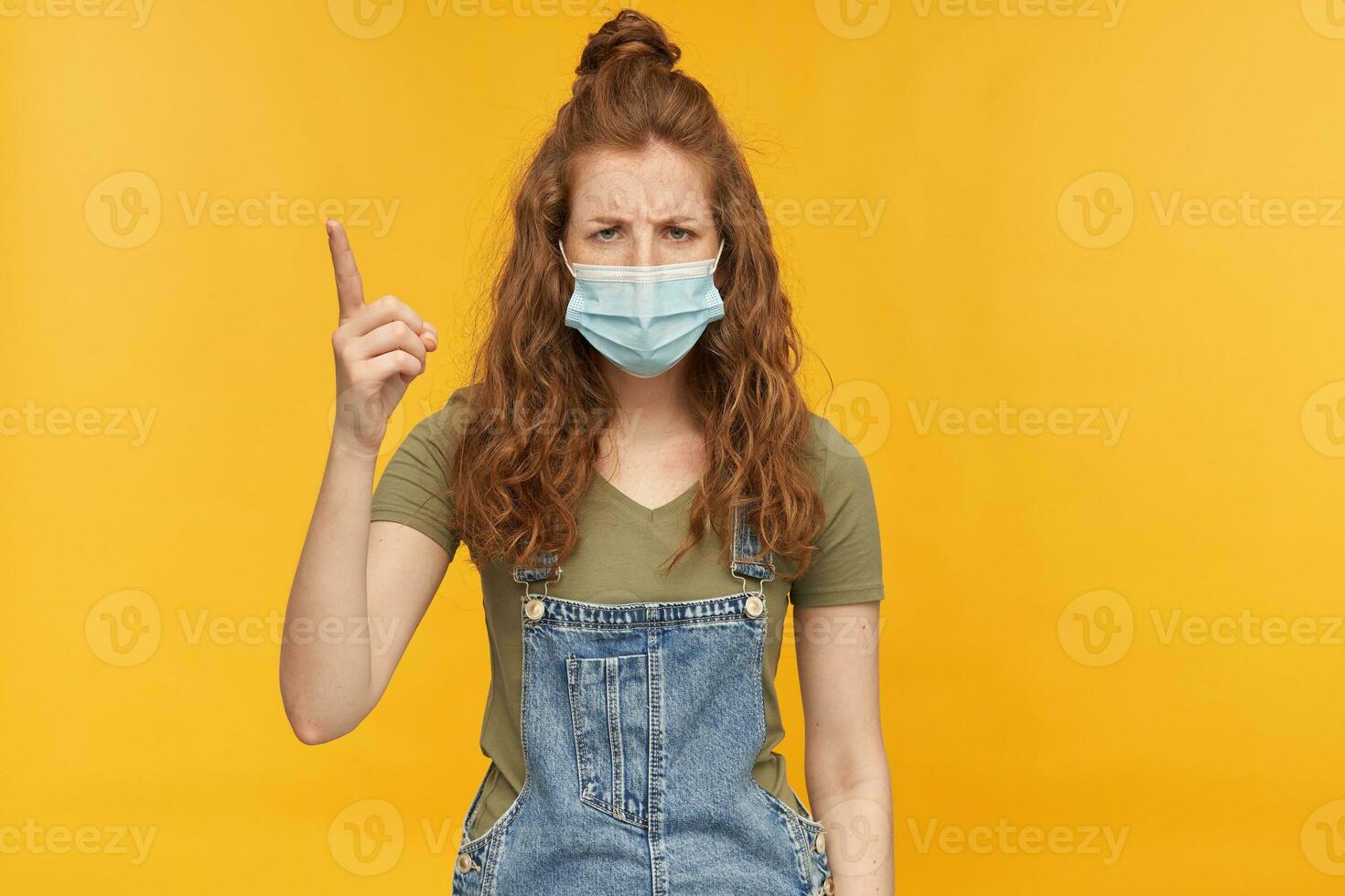indoor studio shot of young ginger female with long red curly hair wears face mask, raised her hand with angry facial expression. isolated over yellow background photo