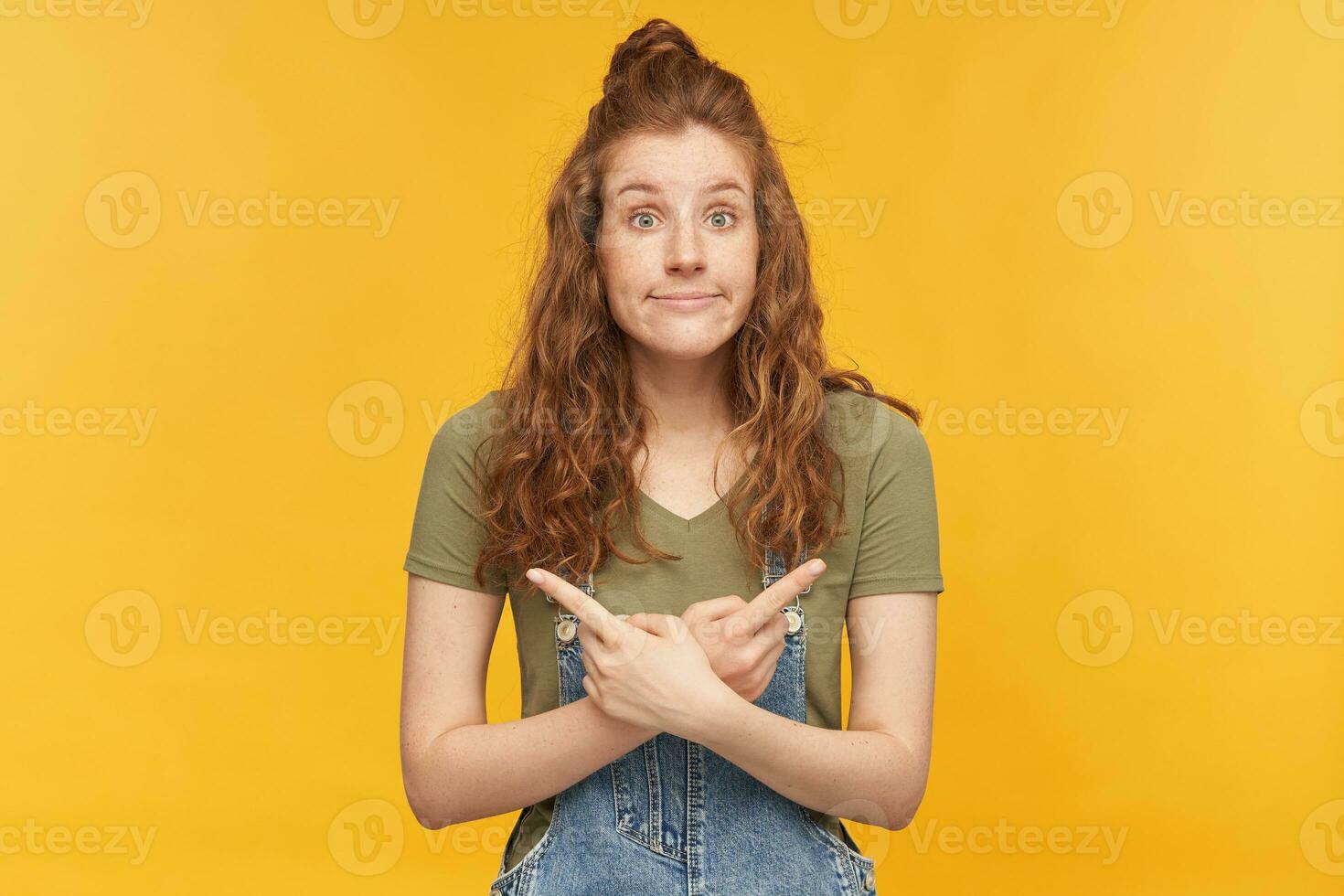 indoor portrait of young ginger female with a wavy long hair pointing both sides with a fingers with confused, irritated facial expression. isolated over yellow background. photo