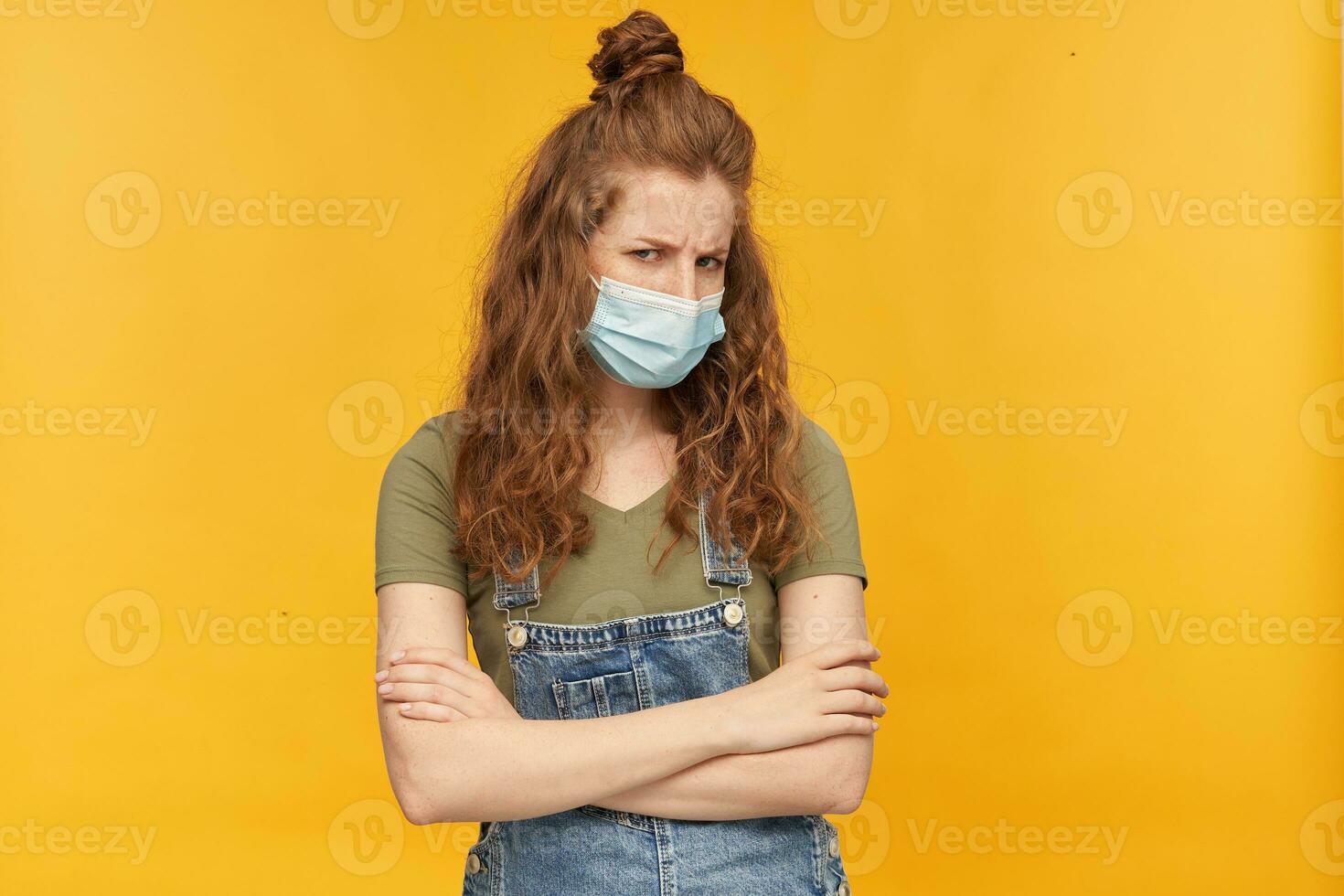 indoor studio shot of young ginger female with long red curly hair looks into camera with angry facial expression, keeps her hands crossed on chest isolated over yellow background photo