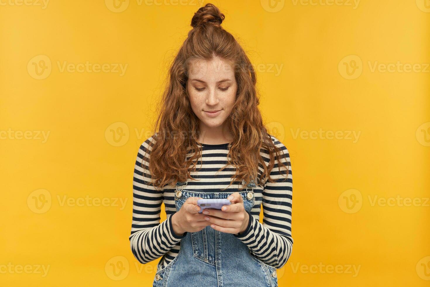 indoor studio shot of young ginger female with long red curly hair keep calm while typing message on her phone isolated over yellow background photo