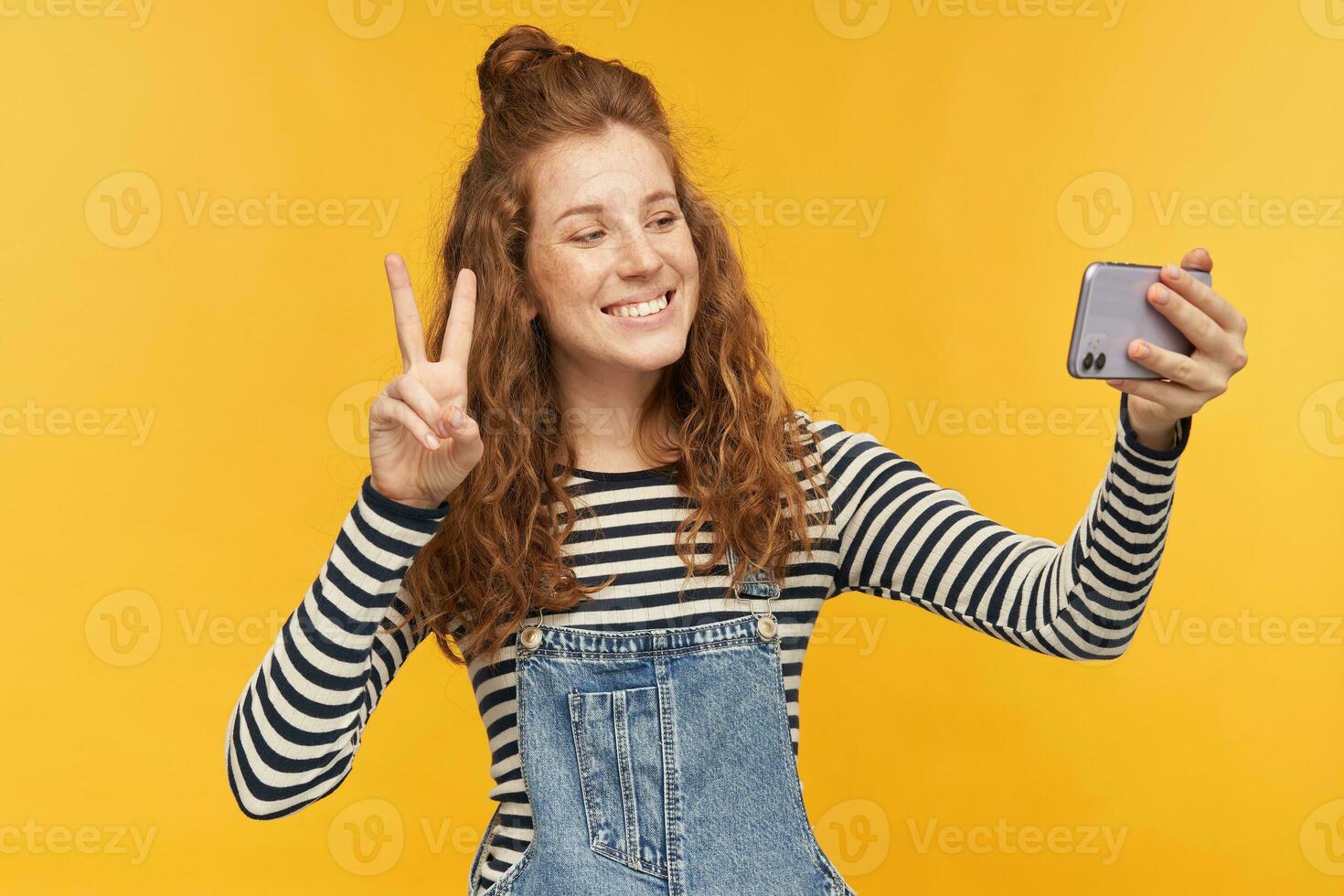 indoor portrait of young happy female, wears stripped shirt and denim overalls shows v sign while have video chat with her boyfriend while isolation. photo