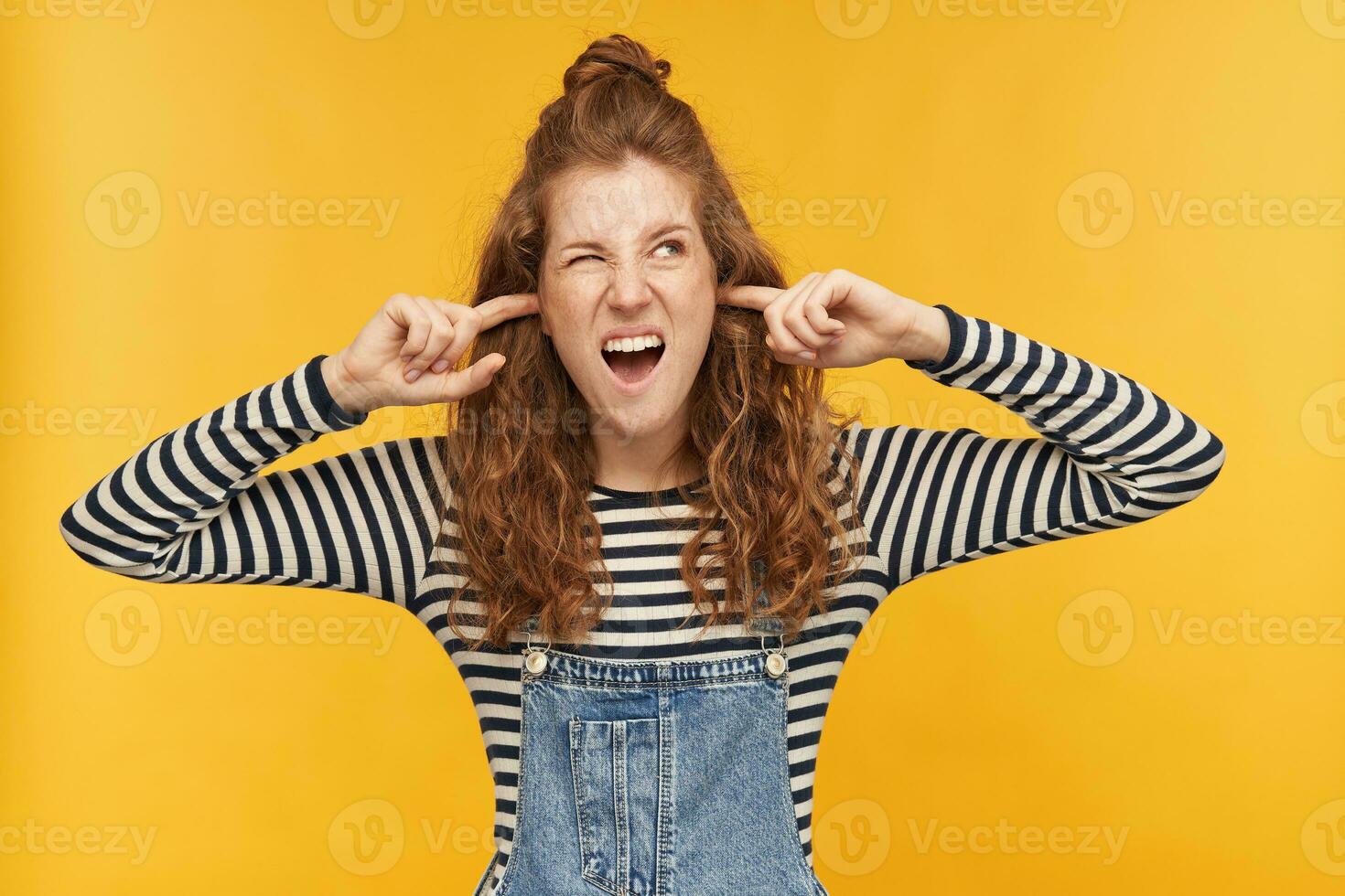Indoor shot of young displeased ginger female, looks aside with negative facial expression, plug her ears with a fingers and mad about noisy neighbors. isolated over yellow background. photo