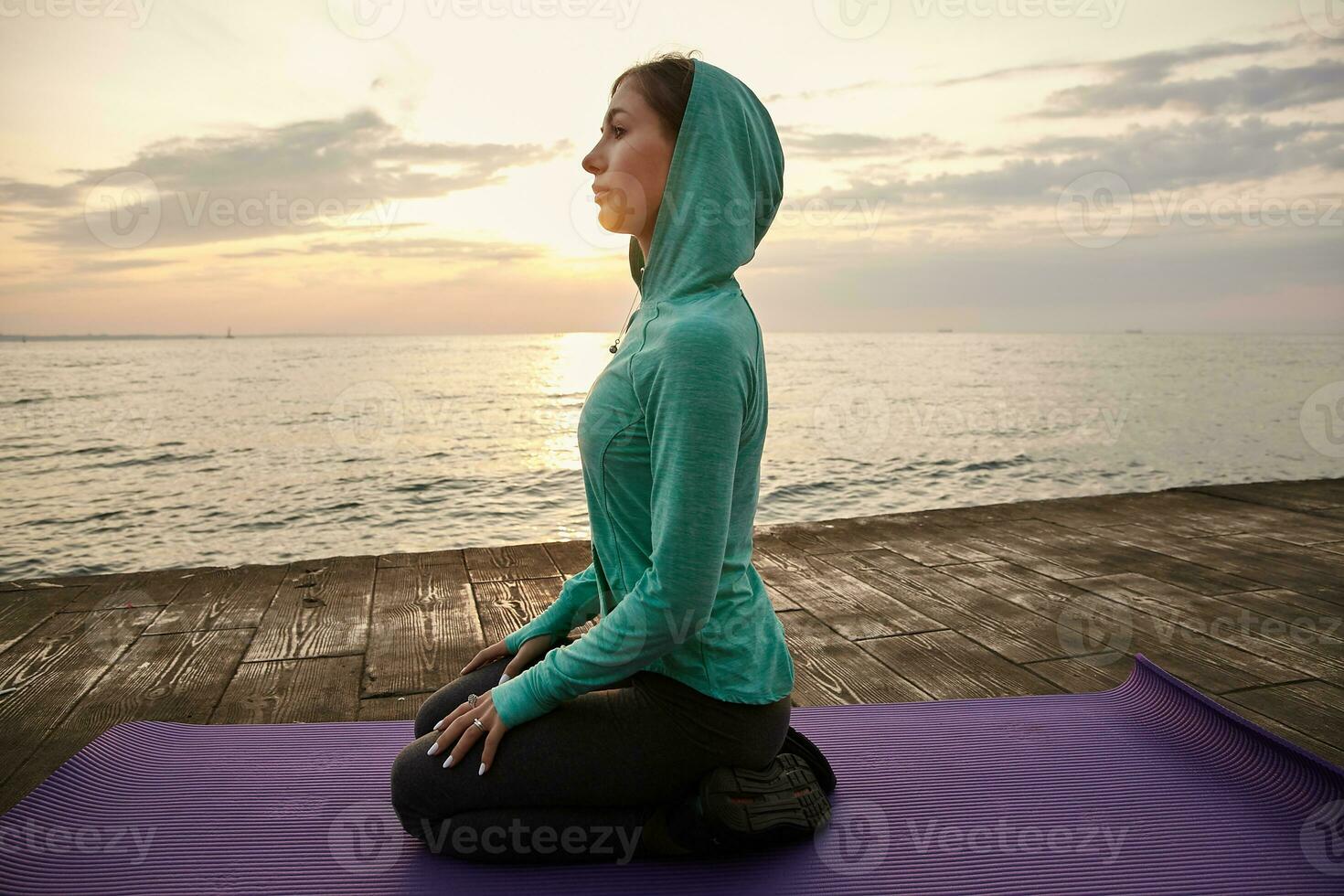 Photo of sporty young woman in yoga pose, dressed in bright sportwears, trains at the seaside, calming and meditate. Health care concept.