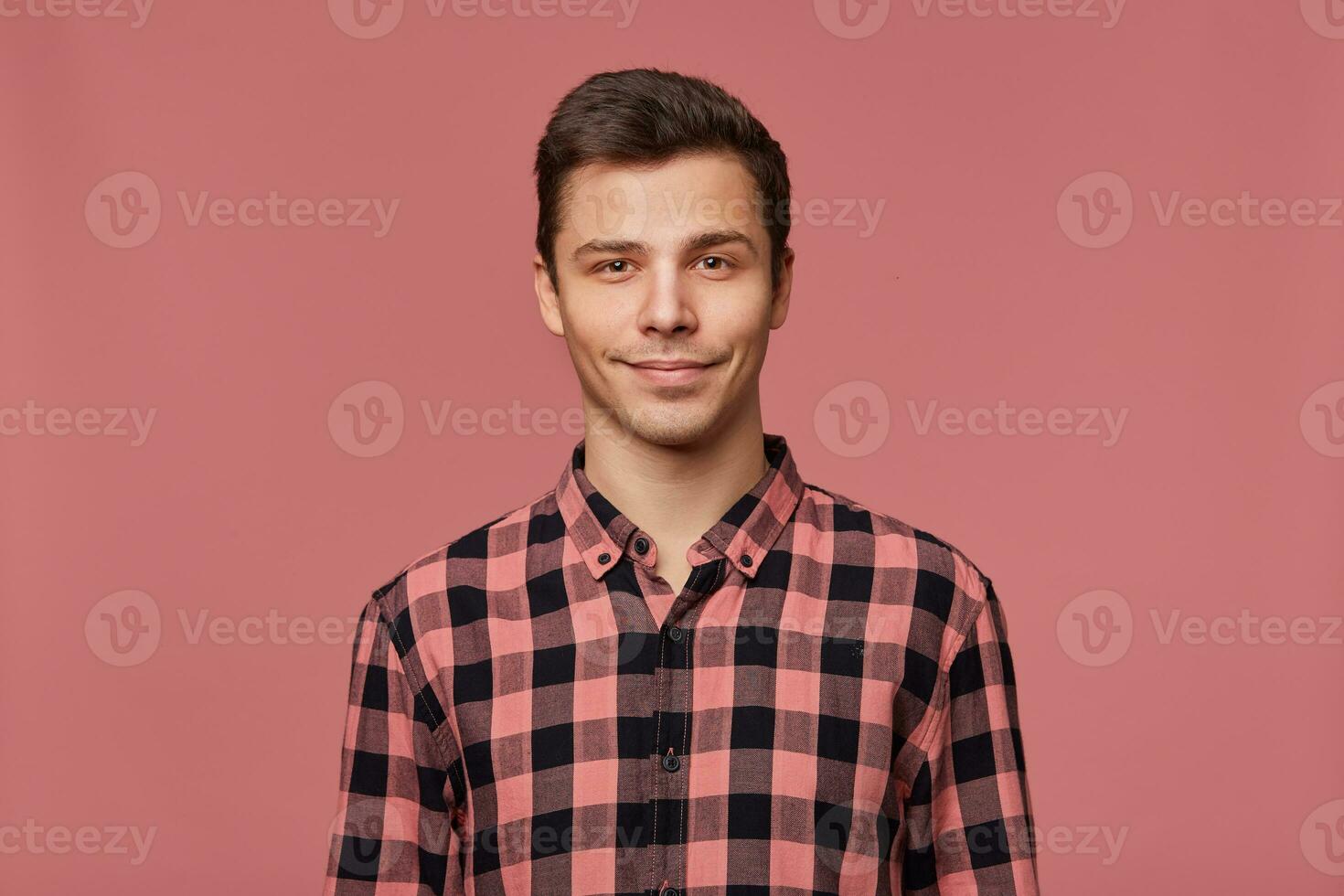 Portrait of young attractive man in checkered shirt, calmly looks at the camera, stands over pink background and smiles. photo