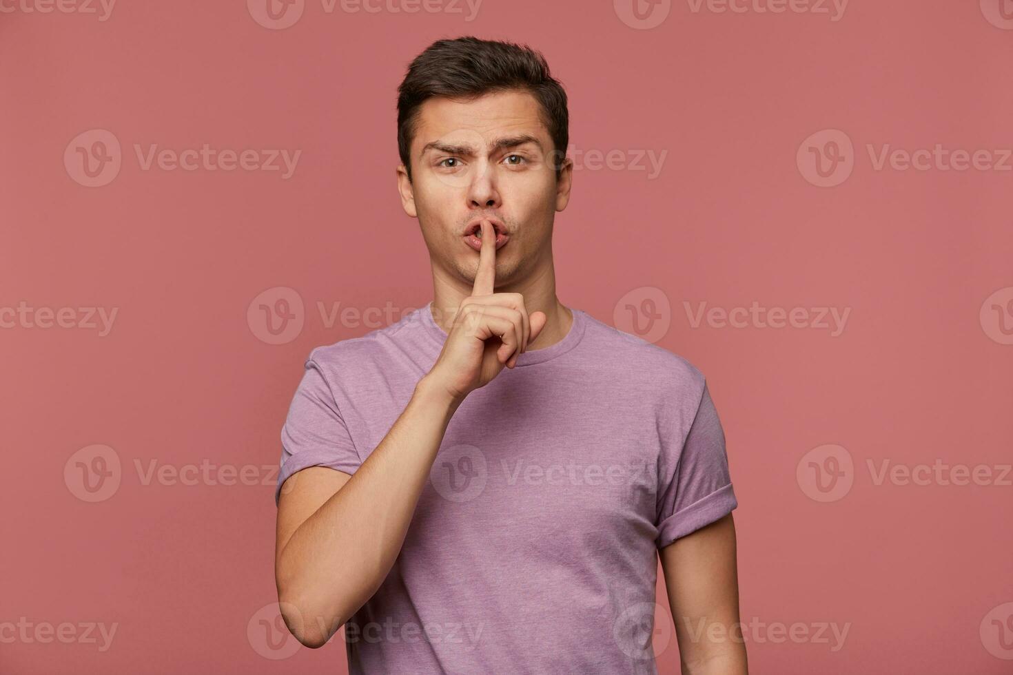 Phortrait of young attractive guy in blank t-shirt, looks at the camera, stands over pink background and shows silence gesture. photo