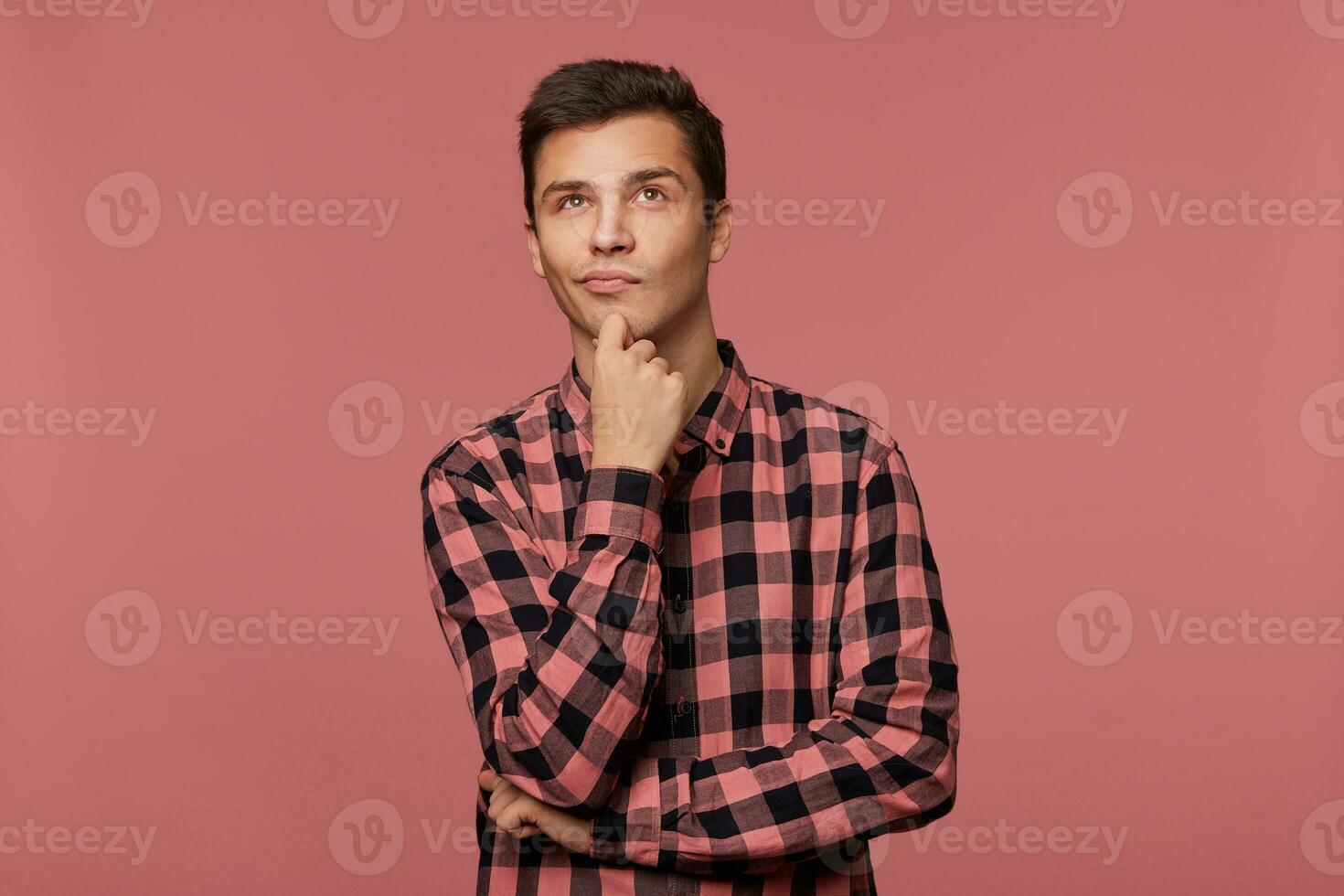 Young handsome pensive guy wears in checkered shirt, looks up with and touches chin, thinks about something, isolated over pink background. photo