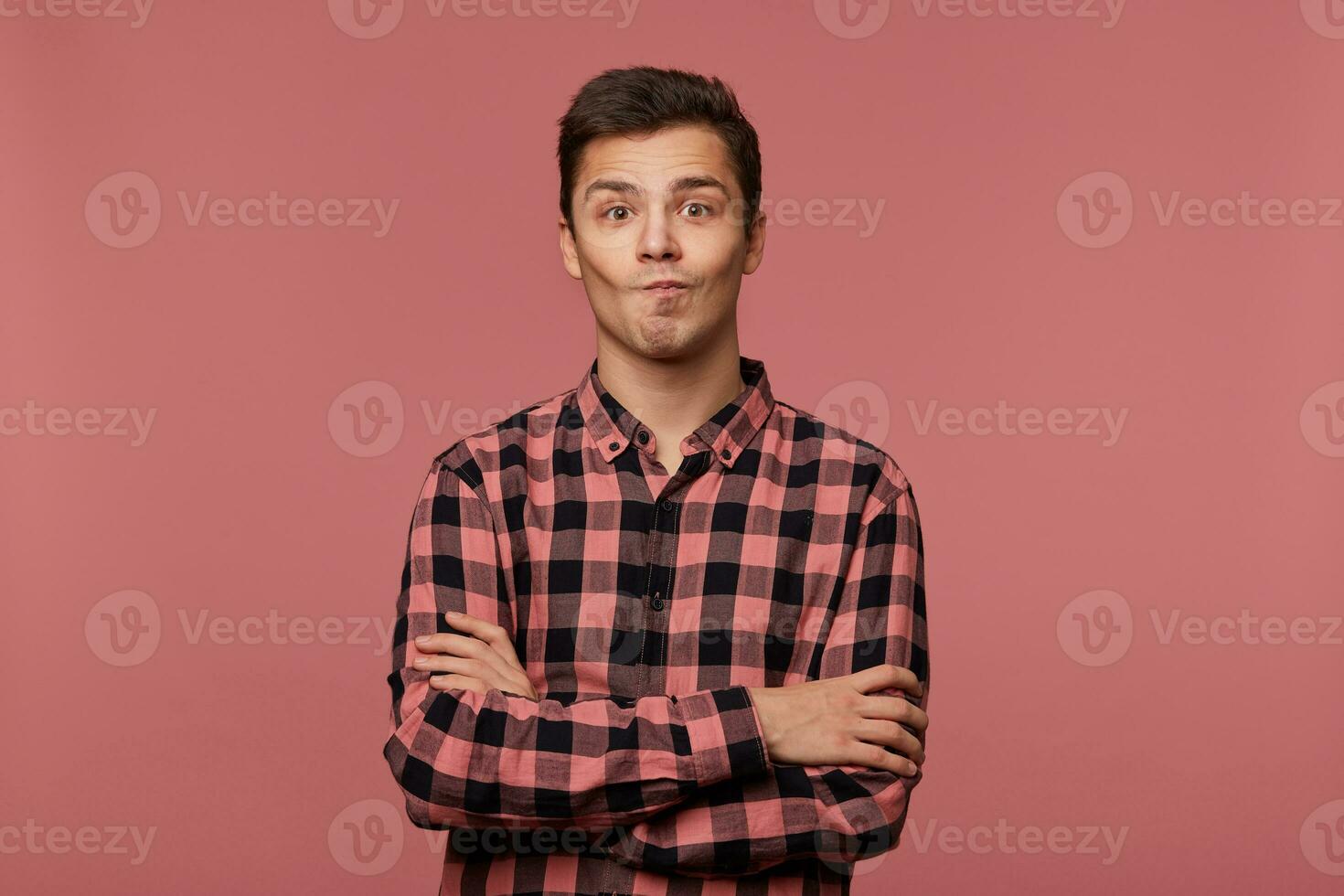 Young handsome wondered guy wears in checkered shirt, looks at the camera with surprised expression, crosed arms, isolated over pink background. photo