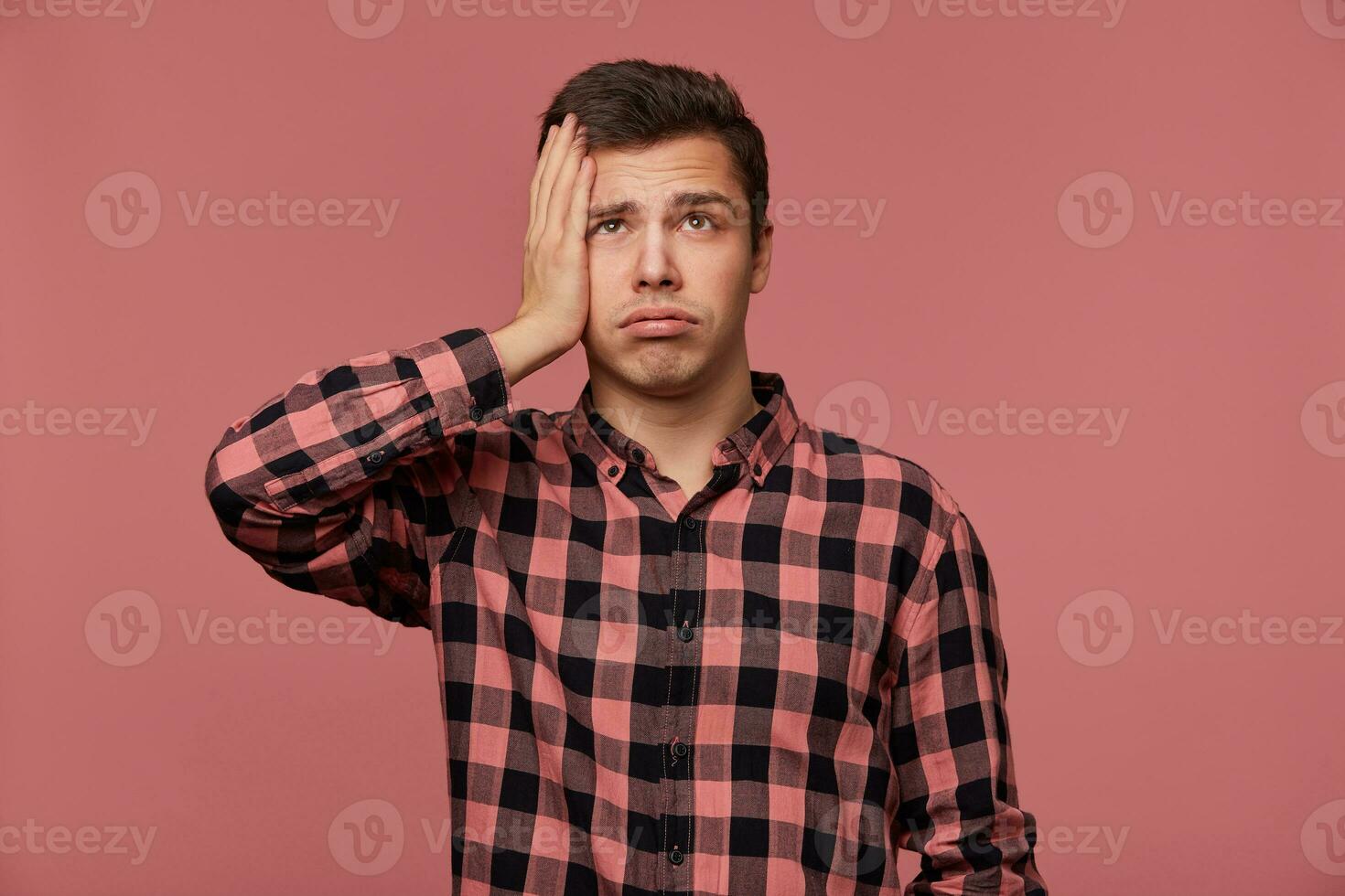 Portrait of young attractive man in checkered shirt, looks up, stands over pink background and touches head, looks tired and sad. photo