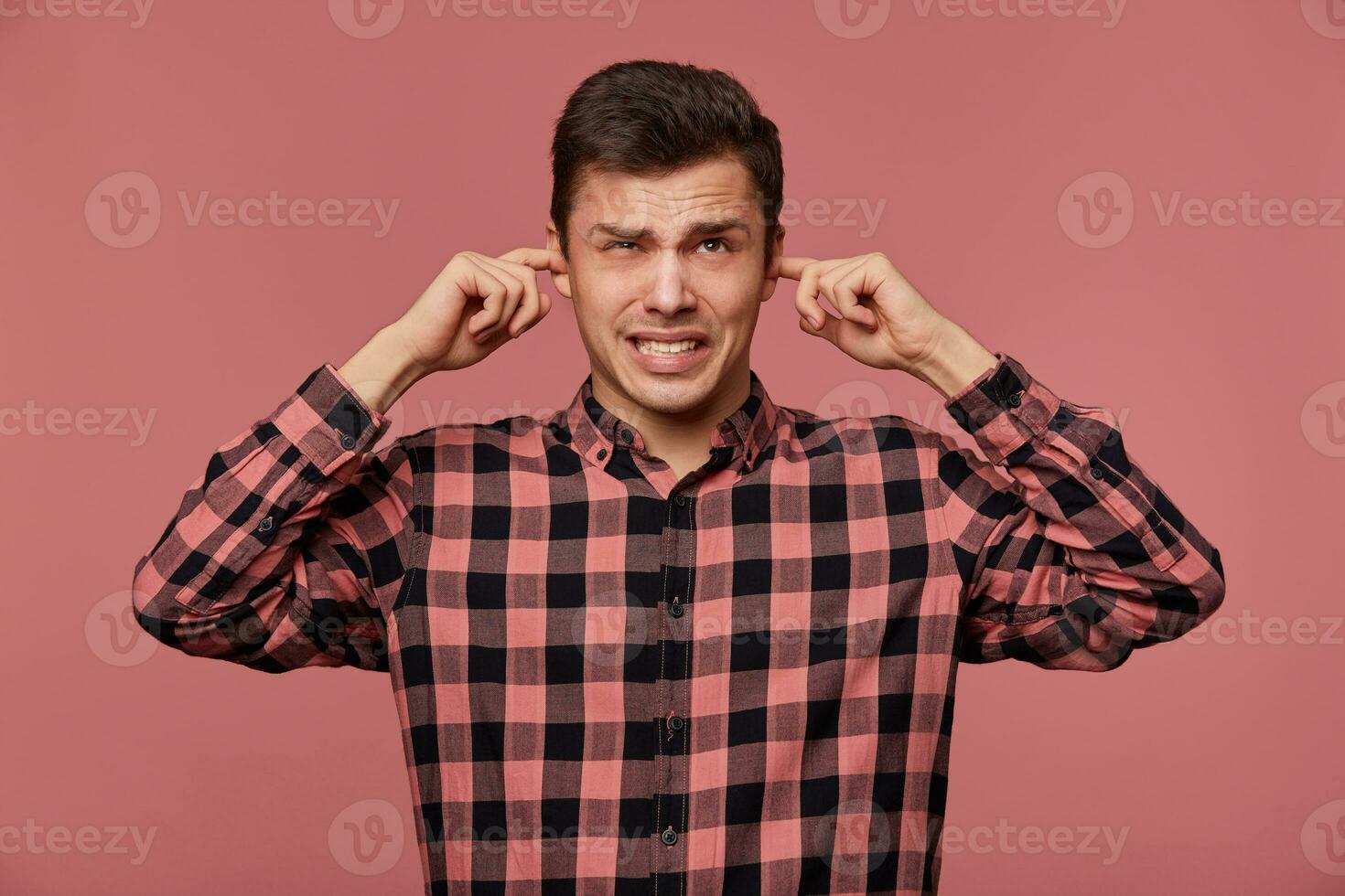 Portrait of young attractive guy in checkered shirt, looks at the camera, stands over pink background and covers ears from loud noise. photo