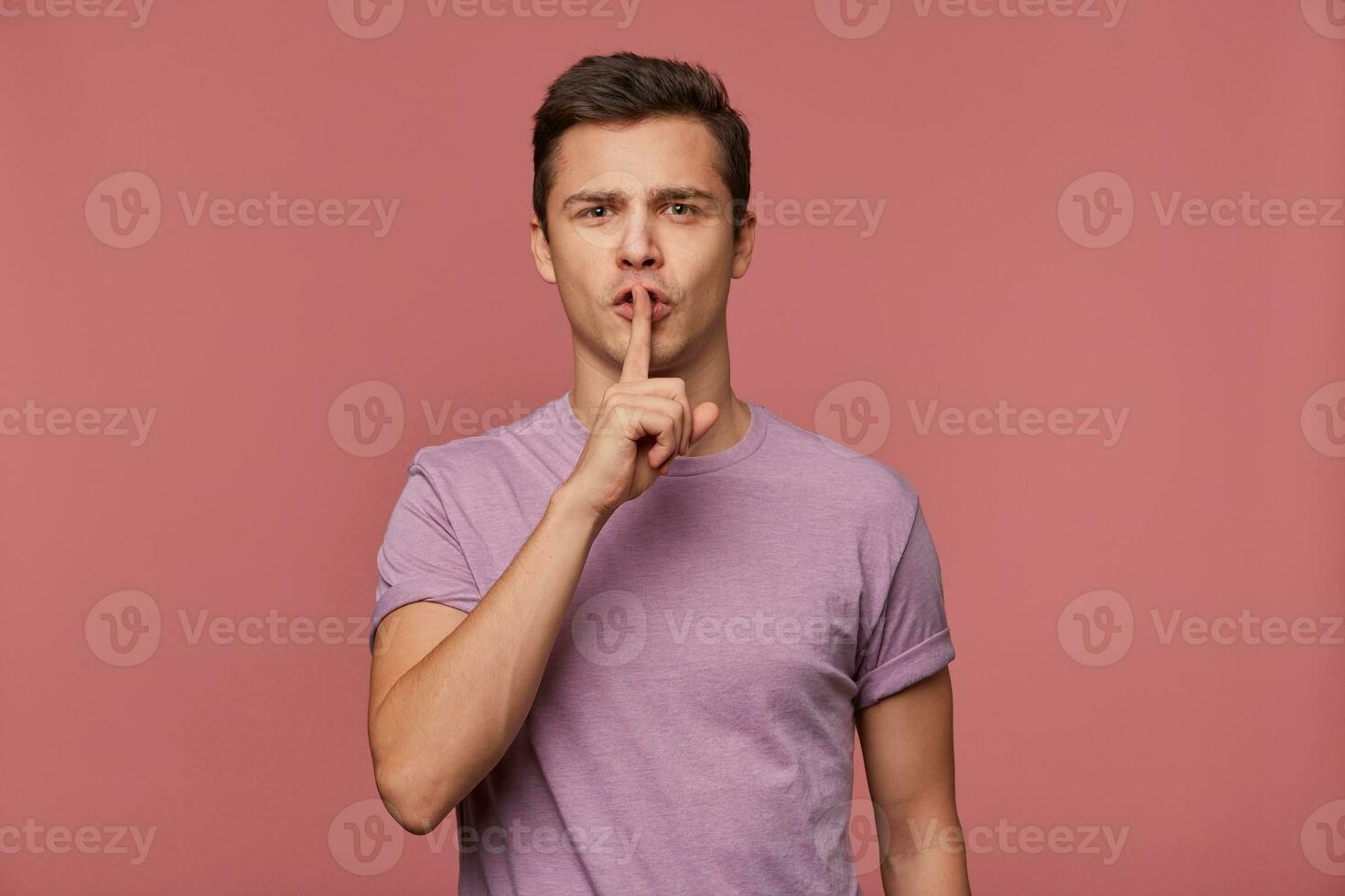 Photo of young attractive man in blank t-shirt, looks at the camera, stands over pink background and shows silence gesture.