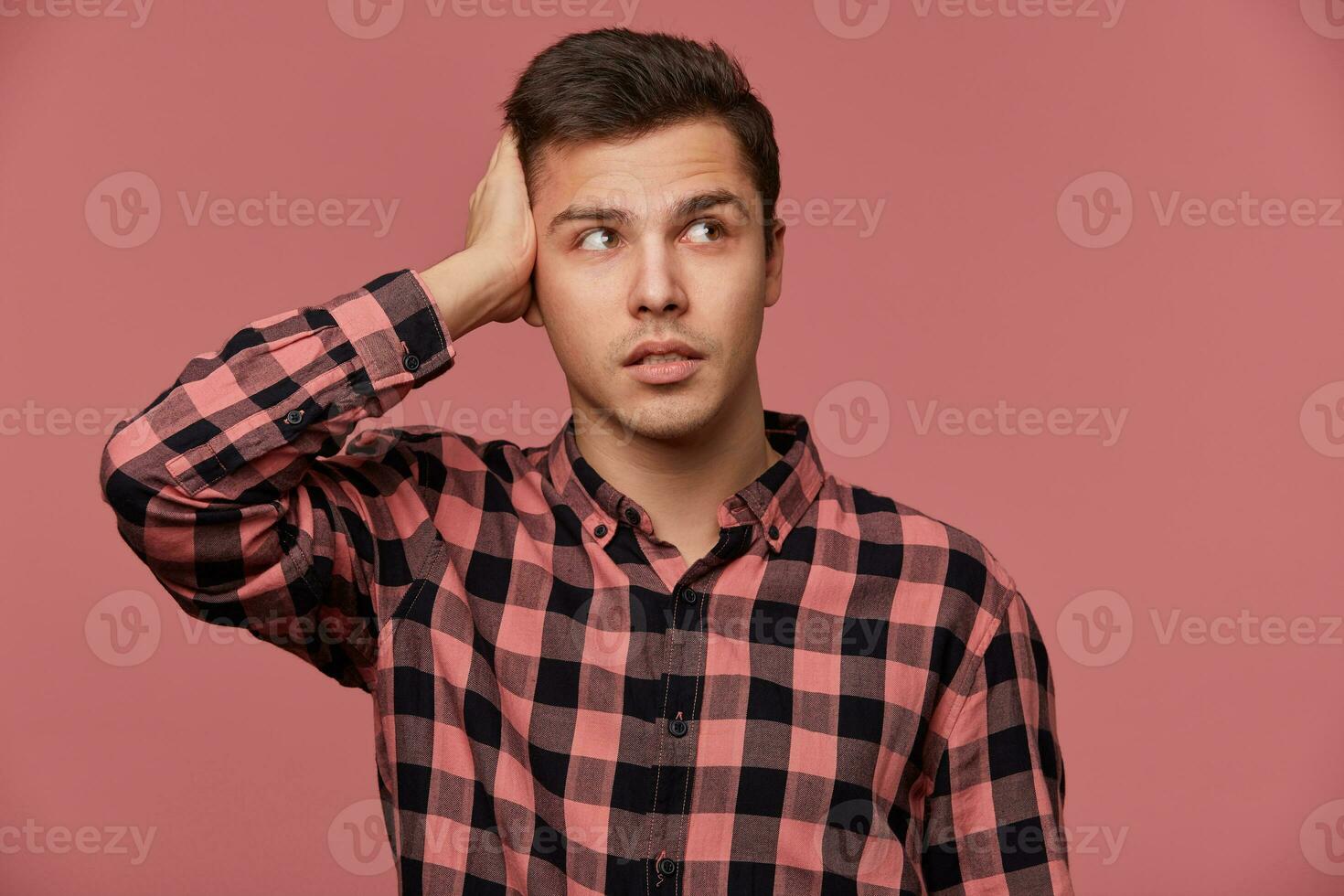 Portrait of young pencive man in checkered shirt, looks away, stands over pink background and touches head, looks tired and sad. photo