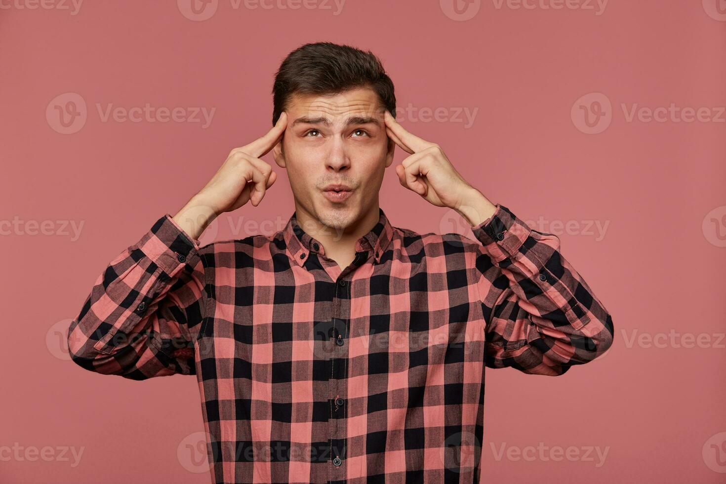 Portrait of young attractive man in checkered shirt, looks at the camera, stands over pink background and shows headshot gesture. photo