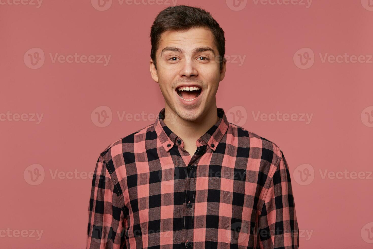 Portrait of young attractive surprised man in checkered shirt, looks at the camera with wide open mouth, stands over pink background. photo