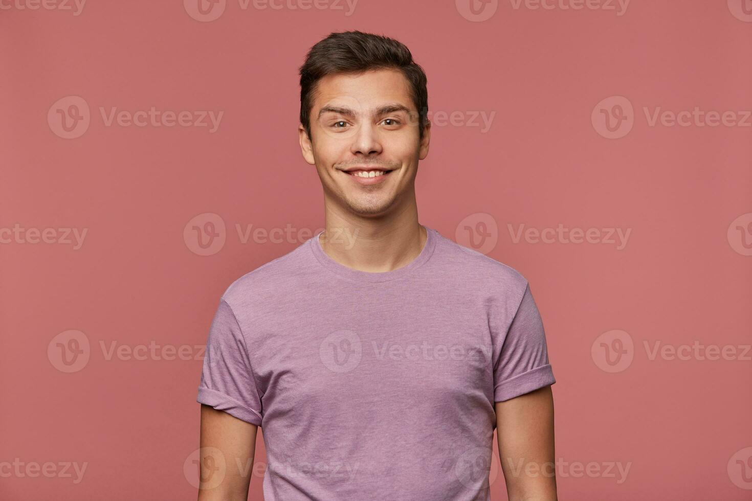 Young handsome cheerful man wears in blank t-shirt, looks at the camera with happy expression, stands over pink background. photo