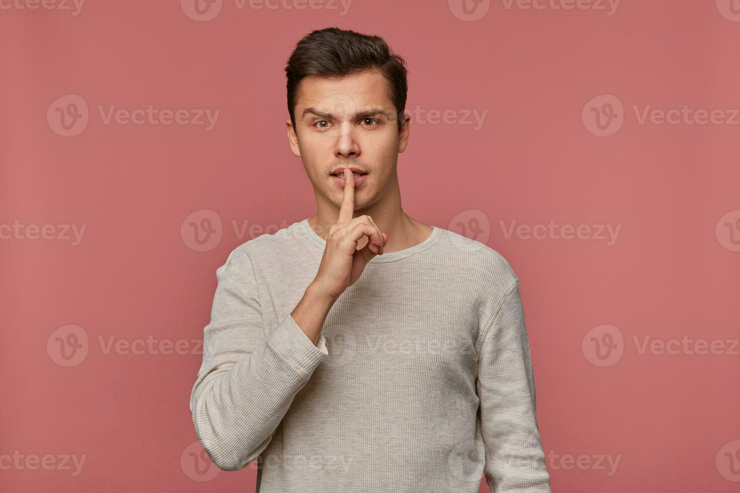 Young attractive man in blank long sleeve, stands over pink background and shows silence gesture, please keep calm and quiet. photo