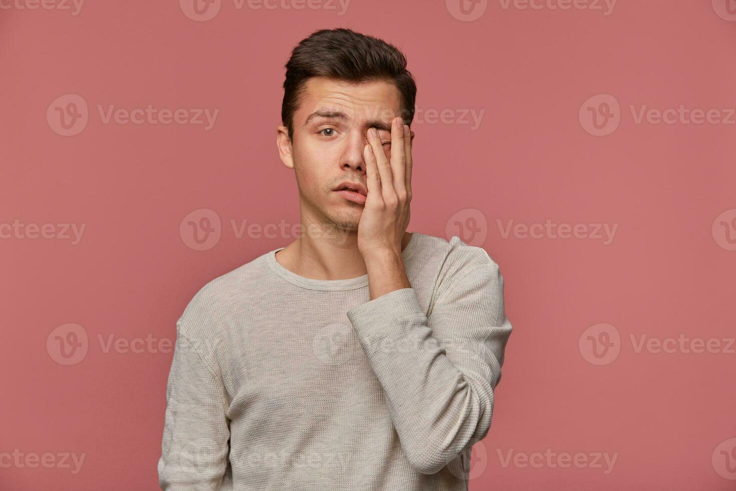 Portrait of young attractive man in white long sleeve, looks at the camera and touches face, stands over pink background, looks tired and sad. photo