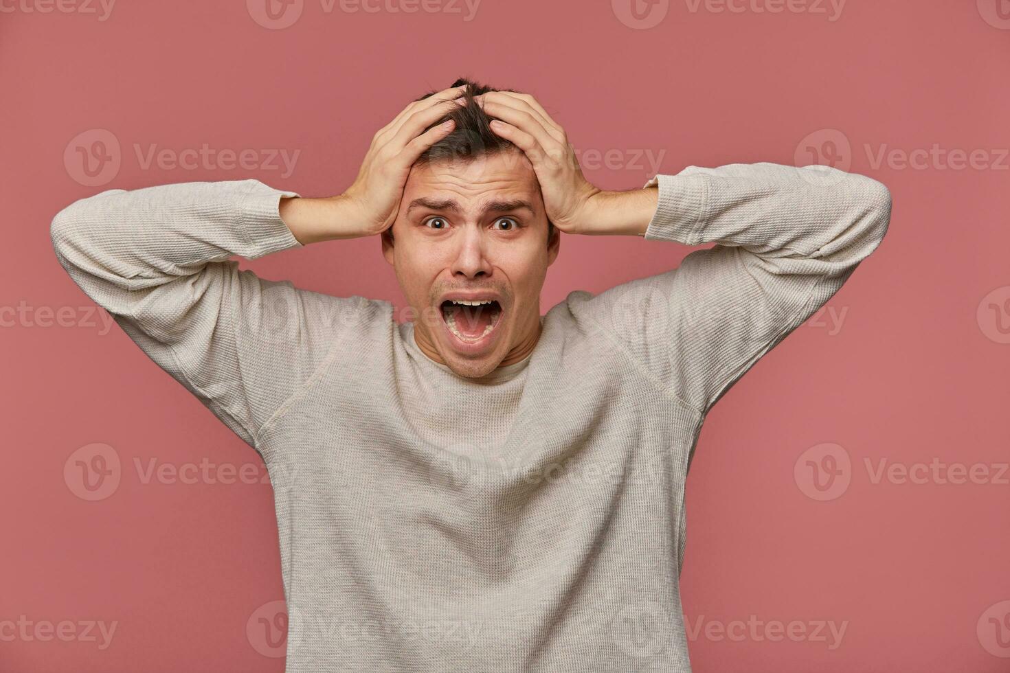 Young scared man in blank long sleeve, hears very bad news, holds head and looks at the camera with shocked expression, stands over pink background and screaming, looks afraid. photo
