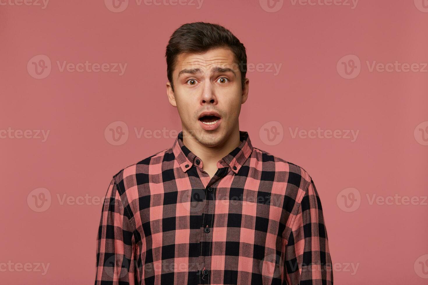 Young handsome amazed man in checkered shirt, looks at the camera with with wide open mouth in surprised expression, isolated over pink background. photo