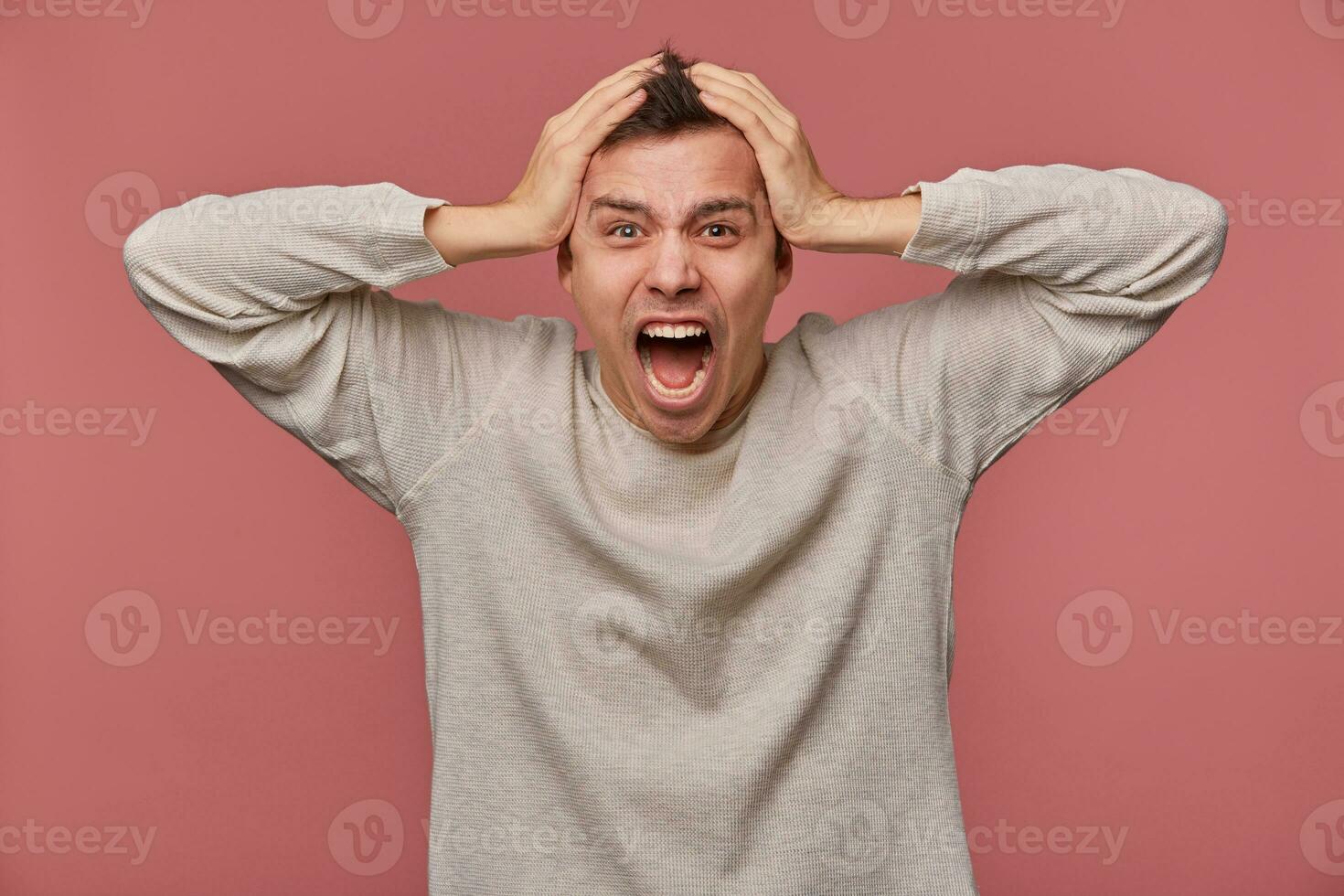 Young scared man in blank long sleeve, holds head and looks at the camera with shocked expression, hears very bad news, stands over pink background and screaming, looks afraid. photo