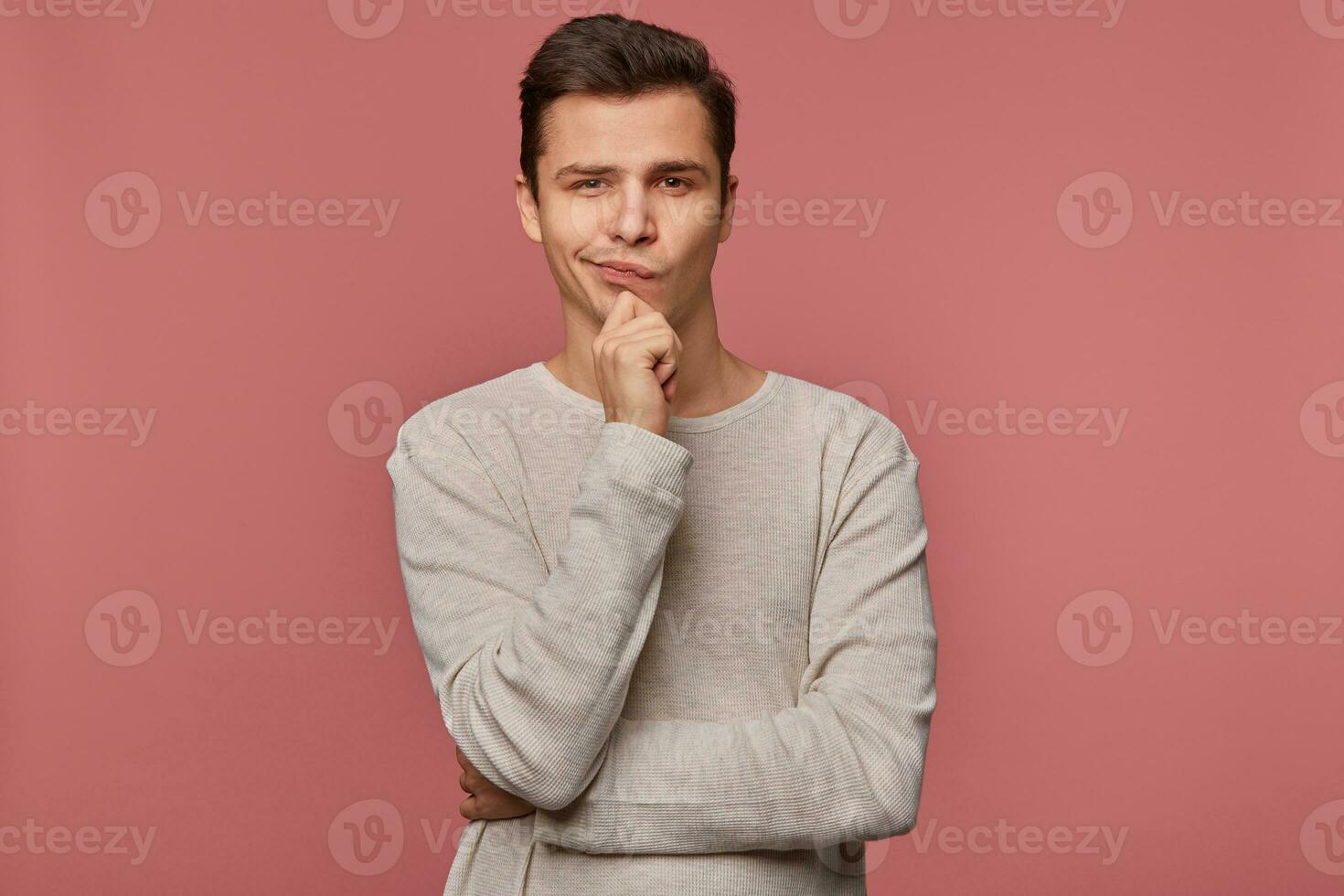 Young handsome thinking guy wears in checkered shirt, looks at the camera and touches chin, thinks about something, isolated over pink background. photo