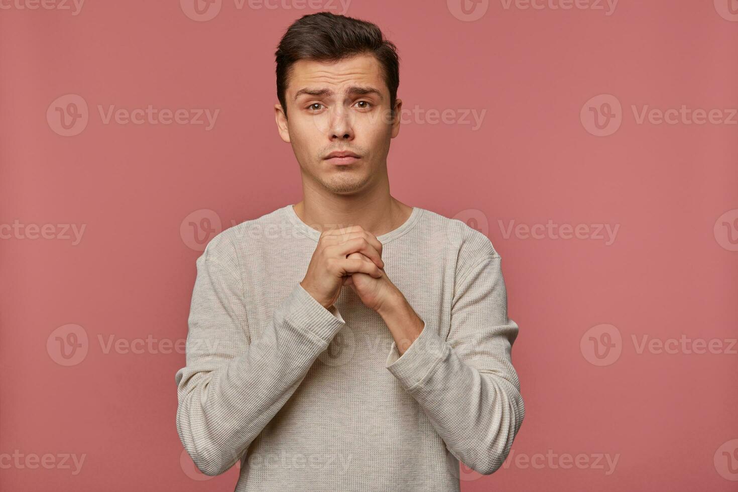 Young attractive guy wears in blank long sleeve, make a wish, hopes for good luck on exams and crossed palms, asks not to give him a bad mark, stands over pink background. photo