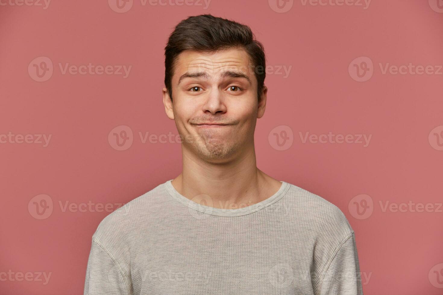 Handsome young sad guy with raised eyebrows wears in blank t-shirt, looks at the camera with mistrust, stands over pink background. photo