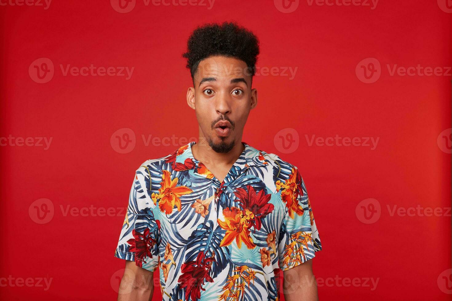 Portrait of shocked young African American guy, wears in Hawaiian shirt, looks at the camera with surprised expression with wide open mouth and eyes, stands over red background. photo