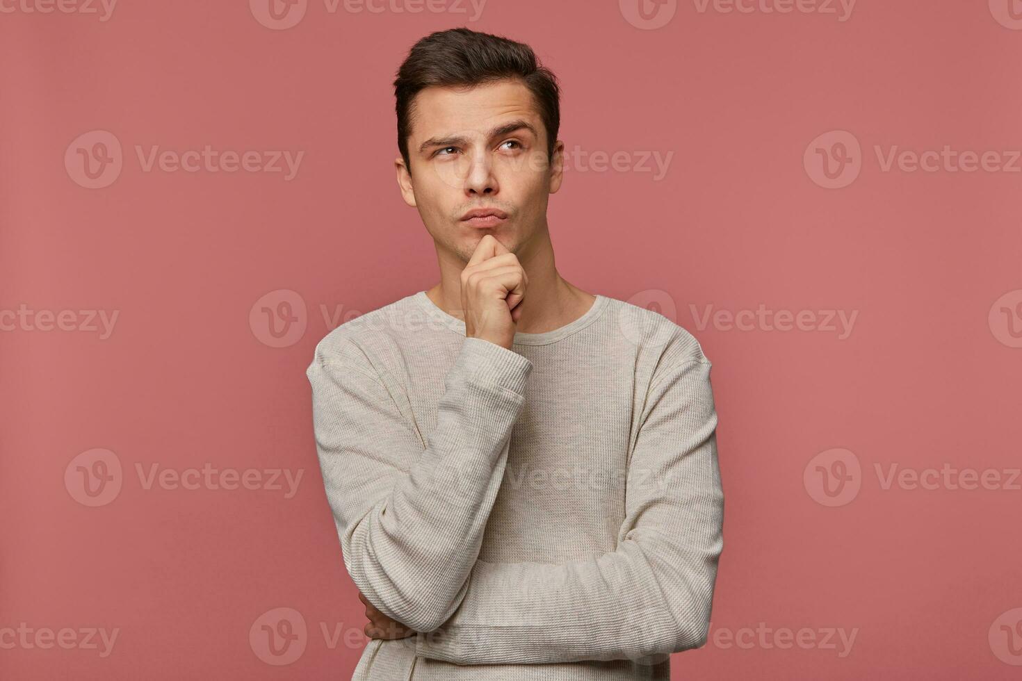 Young handsome thinking guy wears in checkered shirt, looks and away touches chin, thinks about new car, isolated over pink background. photo