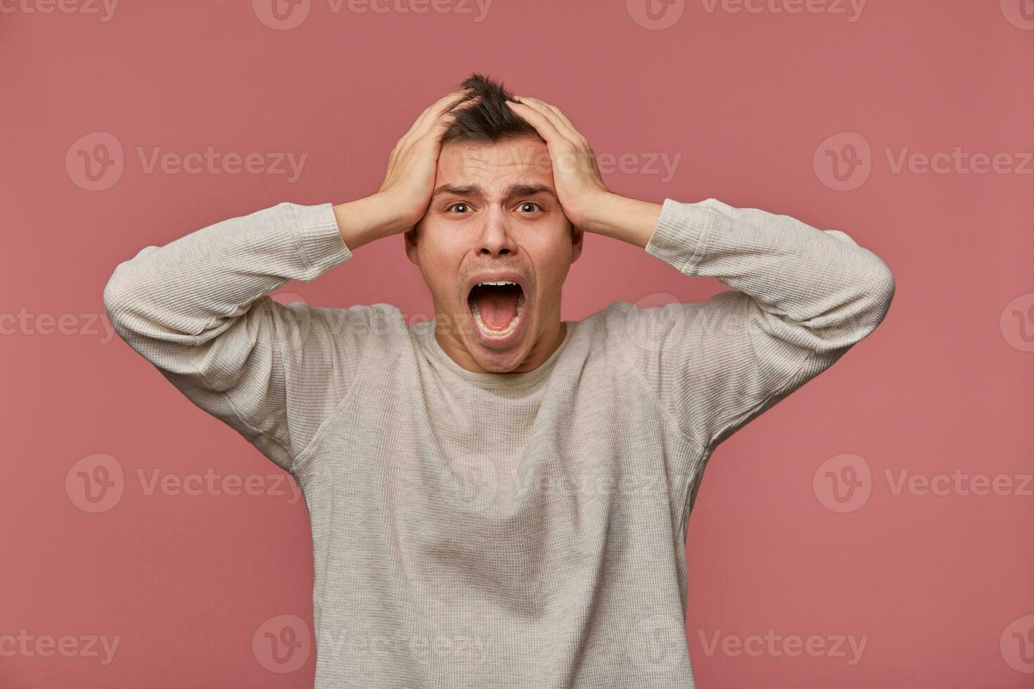 Young scared man in blank long sleeve, holds head and looks at the camera with shocked expression, stands over pink background and screaming, looks afraid. photo