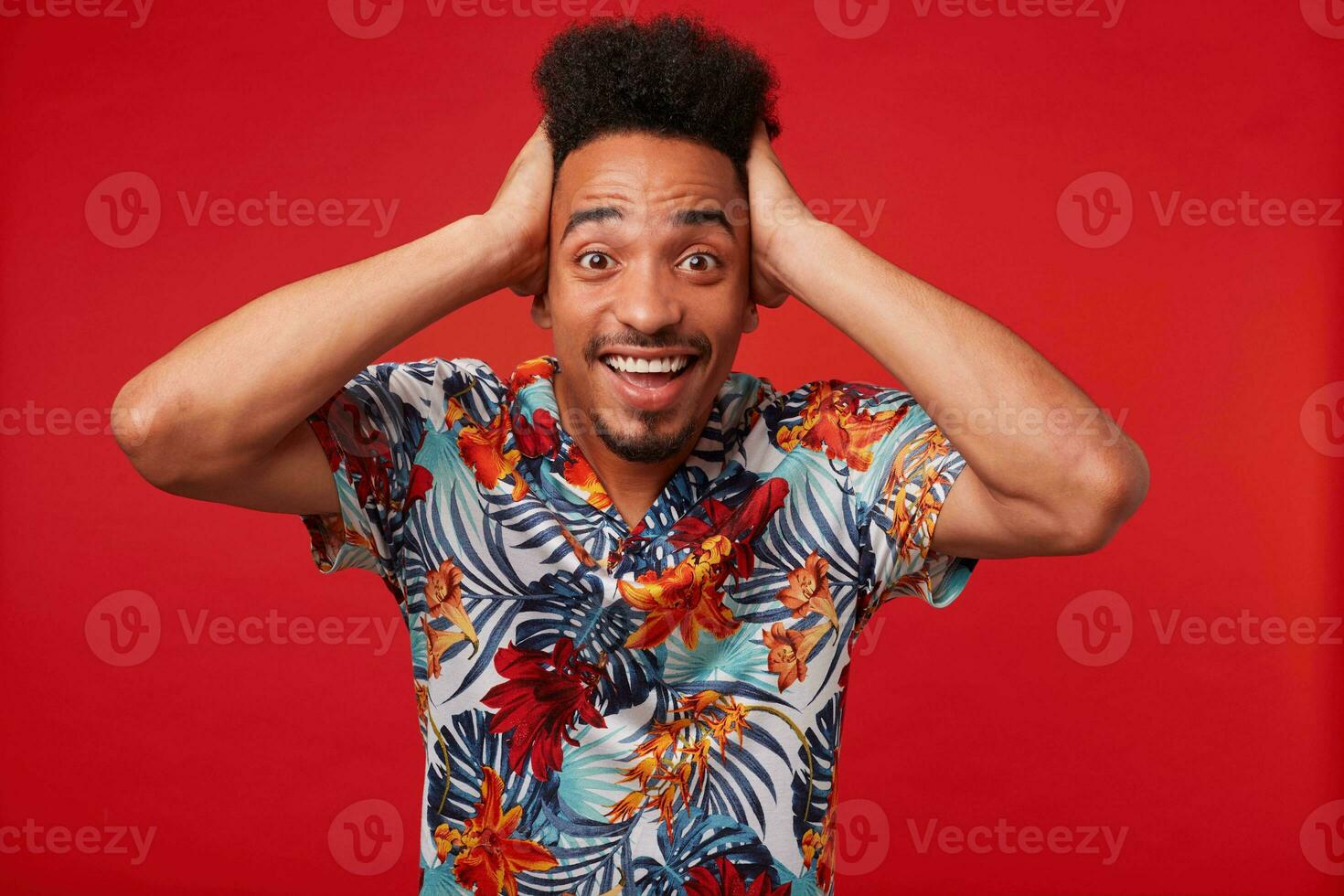 Portrait of young African American man in Hawaiian shirt, looks amazed and happy, stands over red background and holds his head. photo