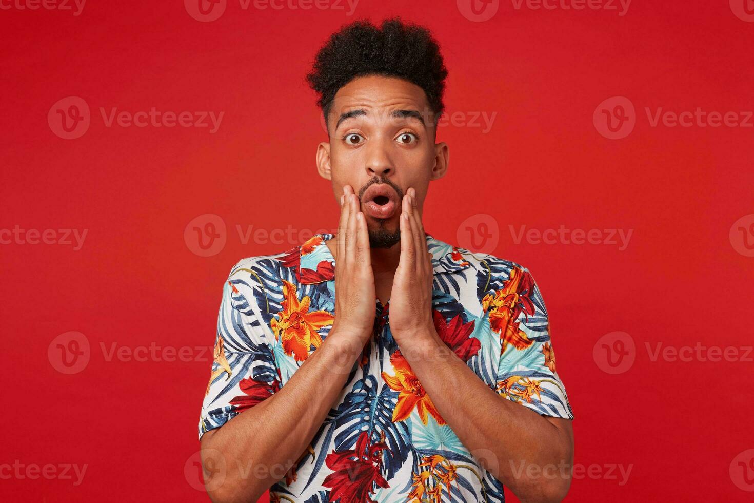 Young shocked African American guy wears in Hawaiian shirt, looks at the camera with surprised expression, stands over red background and touches cheeks. photo