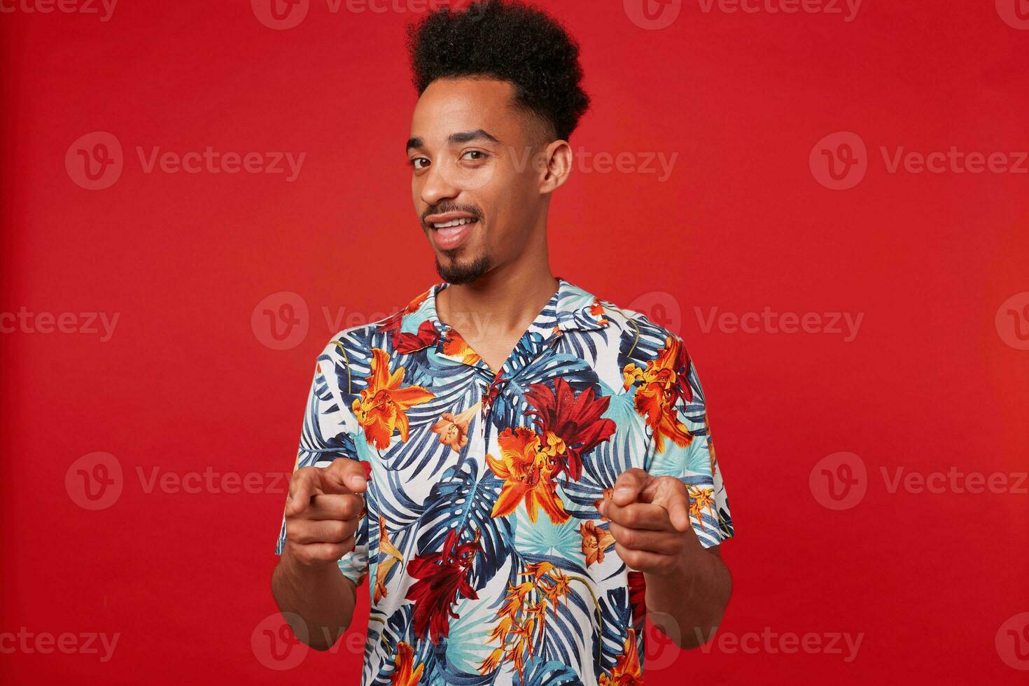 Young positive African American man wears in Hawaiian shirt, looks at the camera and broadly smiles, shows fingers to the camera and says  you cool stands over red background. photo
