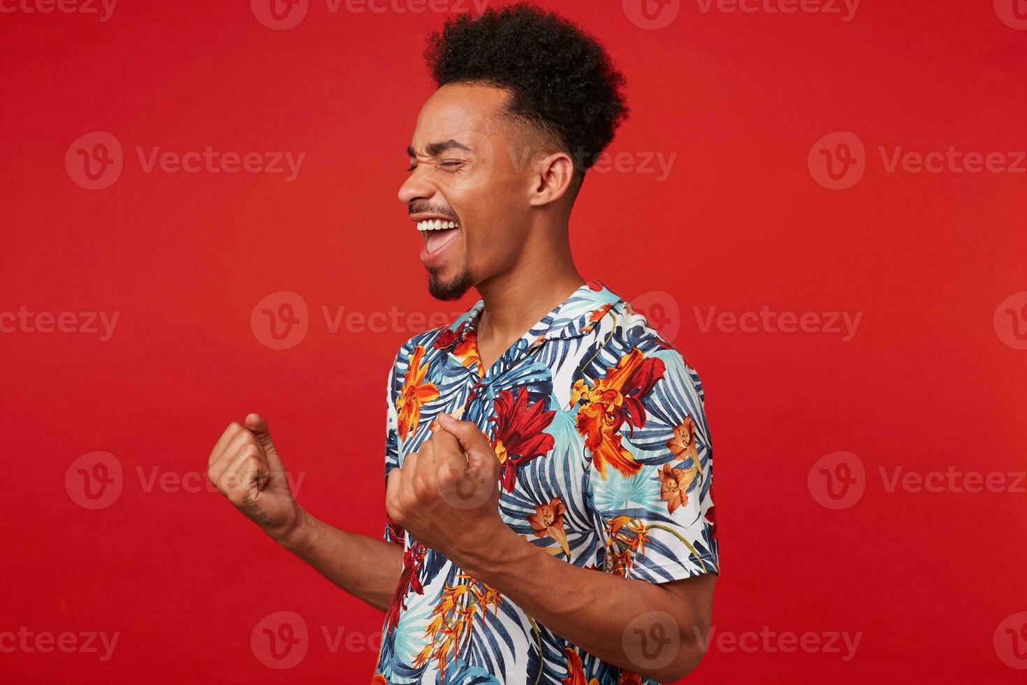 Portrait of young African American man in Hawaiian shirt, looks happy and joyful, stands over red background and clenches fists and rejoices victory. photo