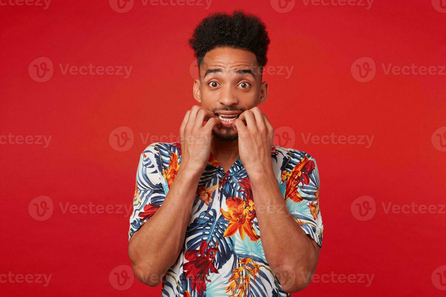 Young scared African American guy wears in Hawaiian shirt, looking at the camera and bites nails, stands over red background. photo