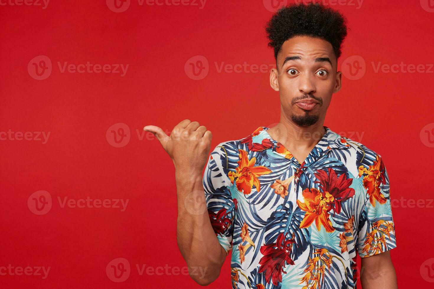 Portrait of young African American man in Hawaiian shirt, looks amazed and happy, stands over red background, points to the left at copyspace. photo