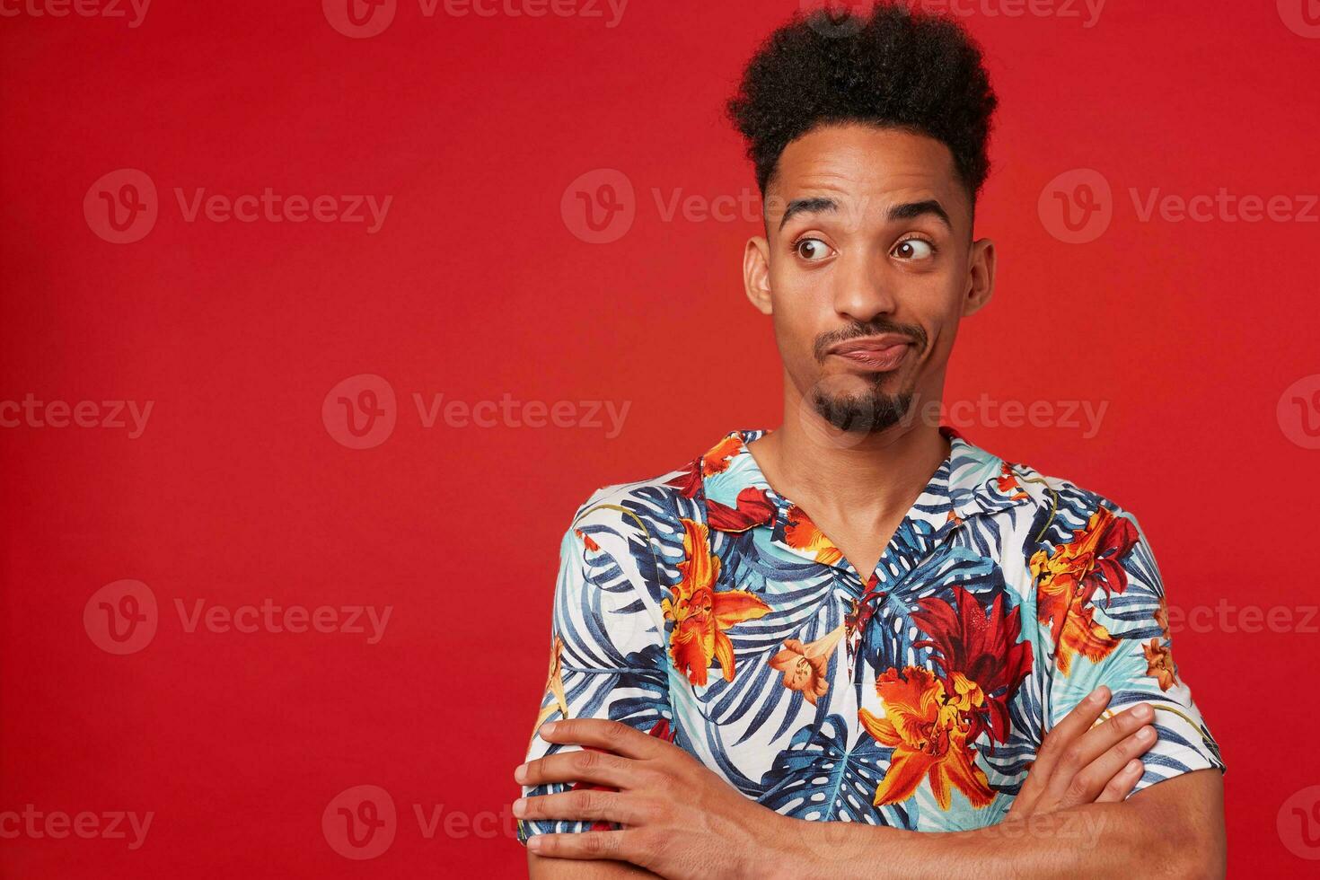 Young unhappy African American guy wears in Hawaiian shirt, looking left at the copy space, stands over red background with crossed arms. photo