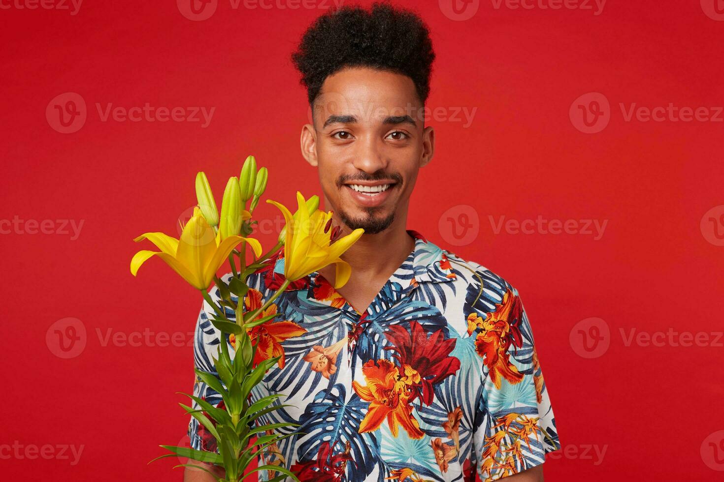 Portrait of cheerful young African American guy, wears in Hawaiian shirt, looks at the camera with happy expression, stands over red background with yellow flowers and broadly smiles. photo
