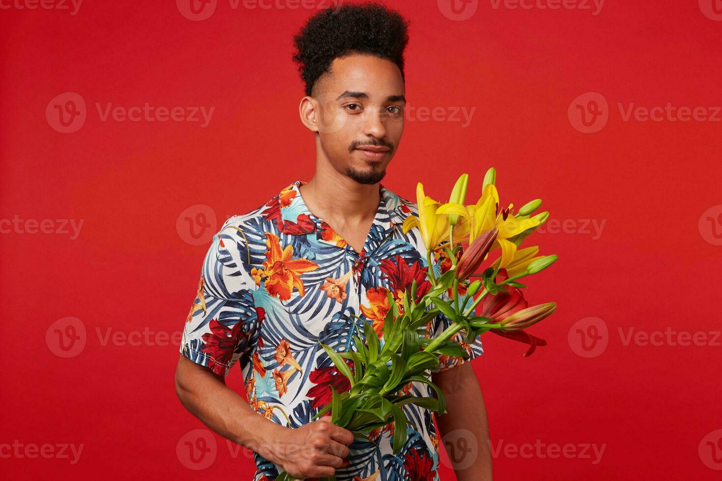 Portrait of cheerful young African American man, wears in Hawaiian shirt, looks at the camera and smile, holds yellow and red flowers, stands over red background. photo