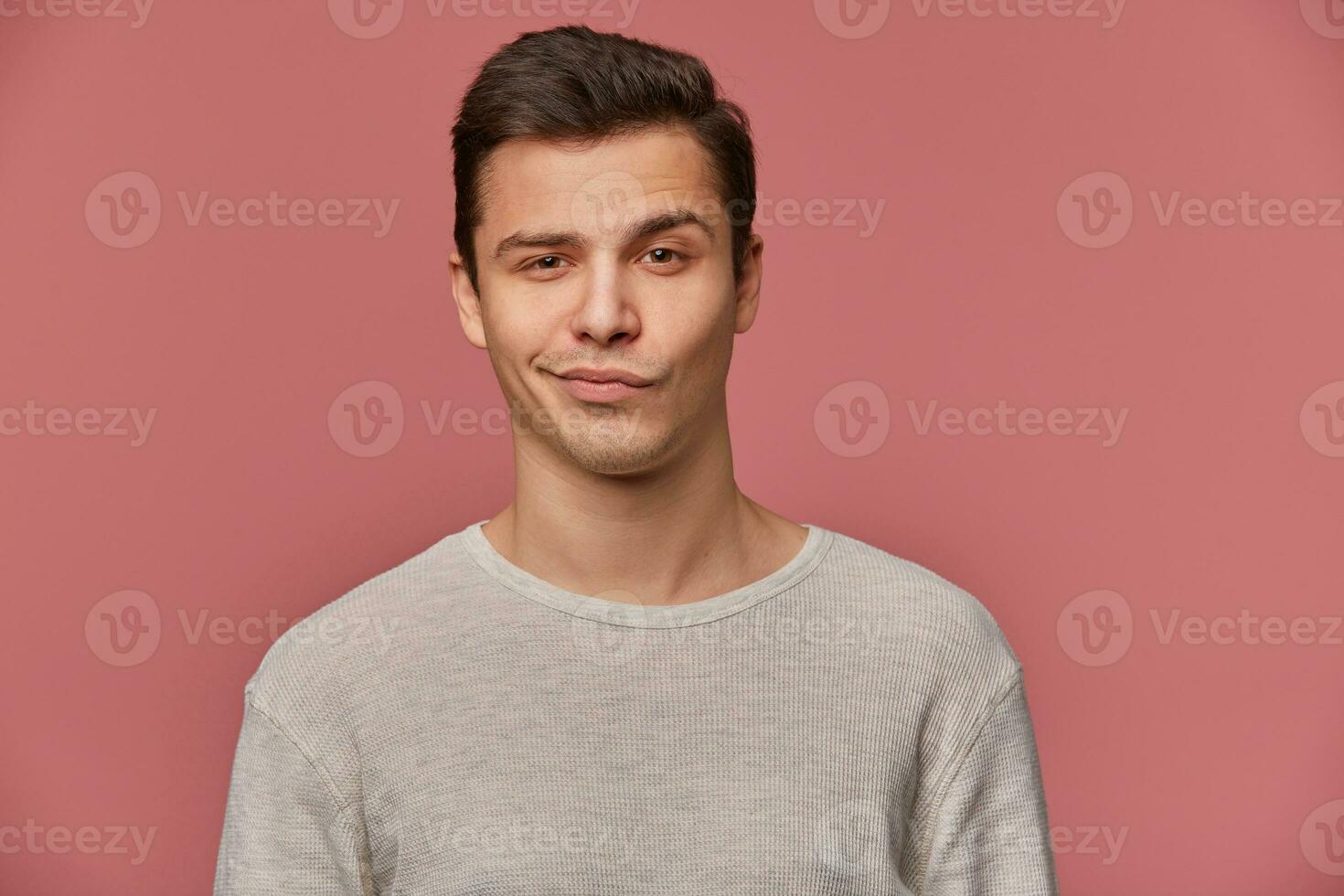 Portrait of handsome young man with raised eyebrow in disapproval, wears in blank t-shirt, looks at the camera with a grin and doubts, stands over pink background. photo
