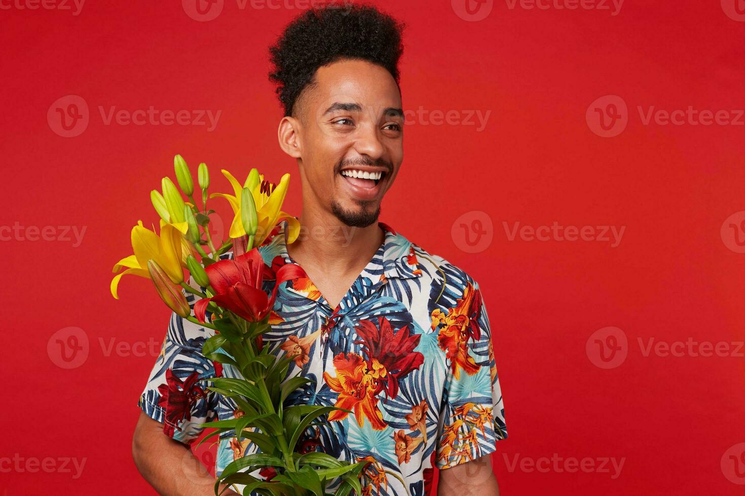 Portrait of laughing young African American guy, wears in Hawaiian shirt, looks at the camera with happy expression, holds yellow and red flowers, stands over red background. photo